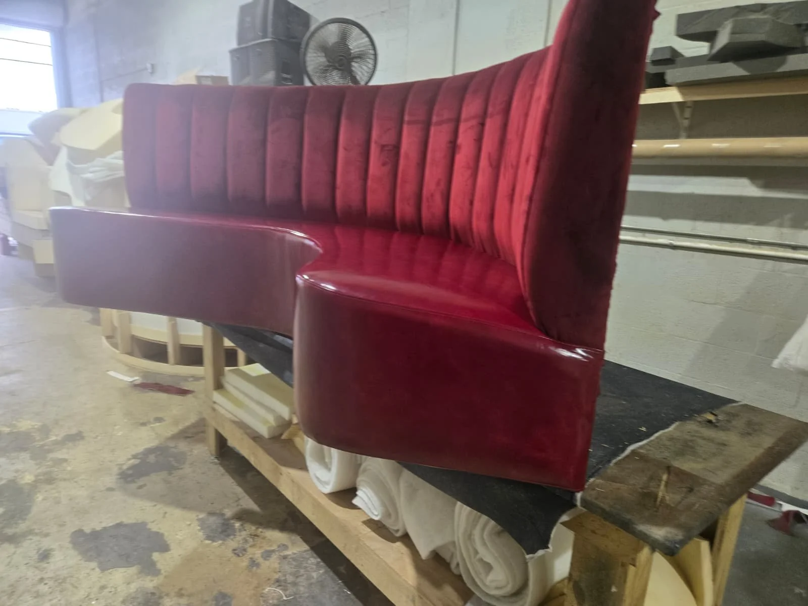 A red upholstered vintage-style booth bench with a curved backrest and armrests, placed on a wooden pallet in a workshop.