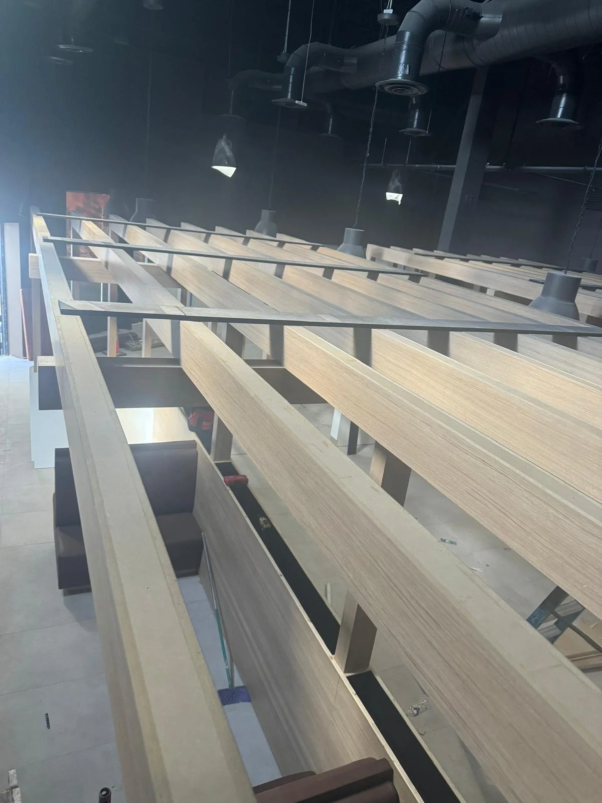 Wooden shelving unit under construction in an industrial-style space with black ceiling pipes and hanging pendant lights.