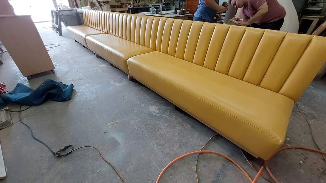 A long yellow upholstered sofa with vertical stitching on the backrest, situated in a workshop or furniture store. The surroundings include various tools, cords, and people working in the background.