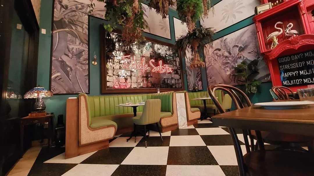 Interior of a restaurant with green booth seating, a small table with place settings, a neon sign on the wall that says 'Fasale Salina,' and decorative plants. There is a red shelving unit with a sign and flamingo figurines on the right, and a checke