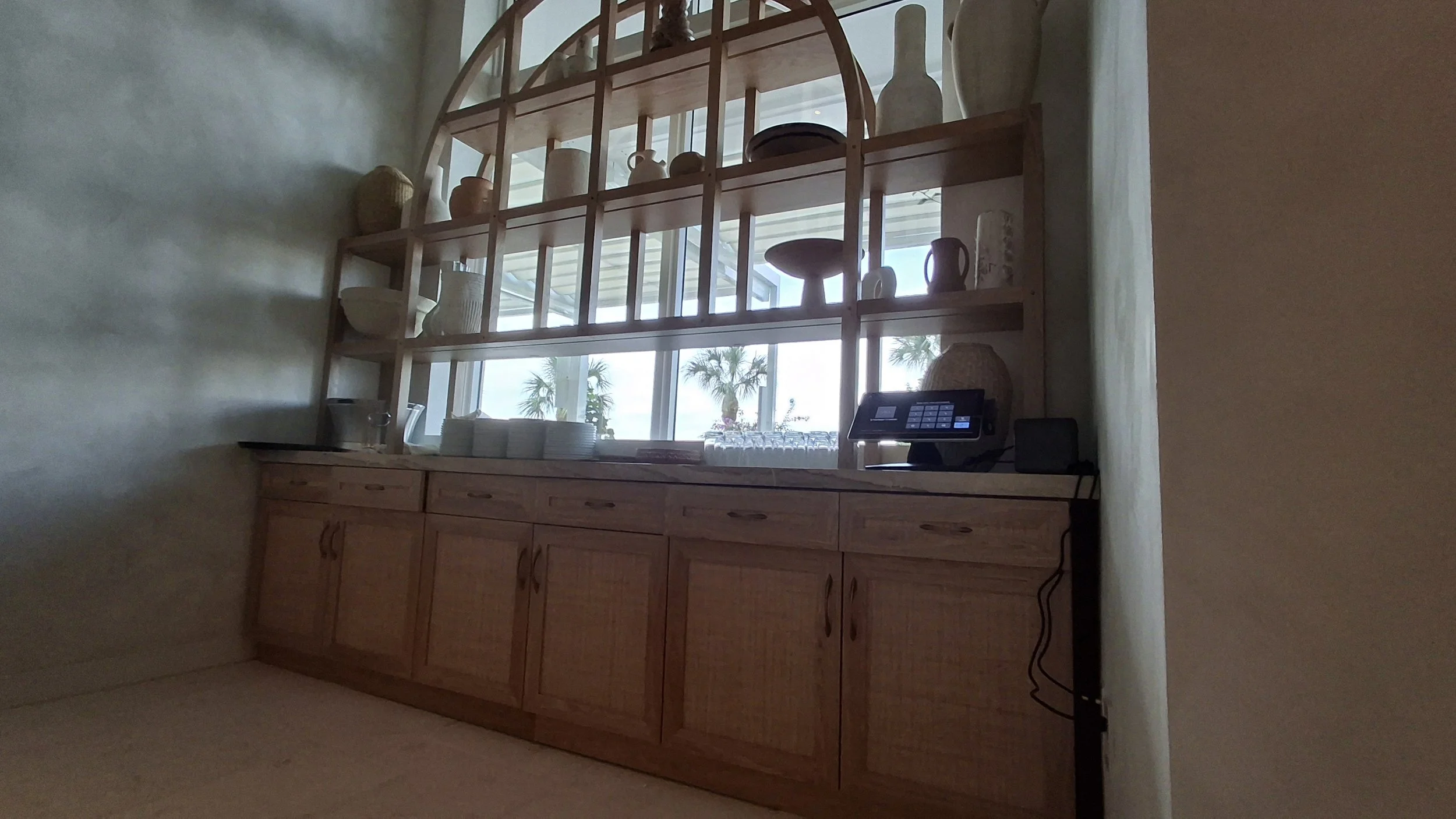 Wooden buffet with drawers and cabinets, topped with plates, bowls, and a point-of-sale terminal, set against a large window with palm trees visible outside.