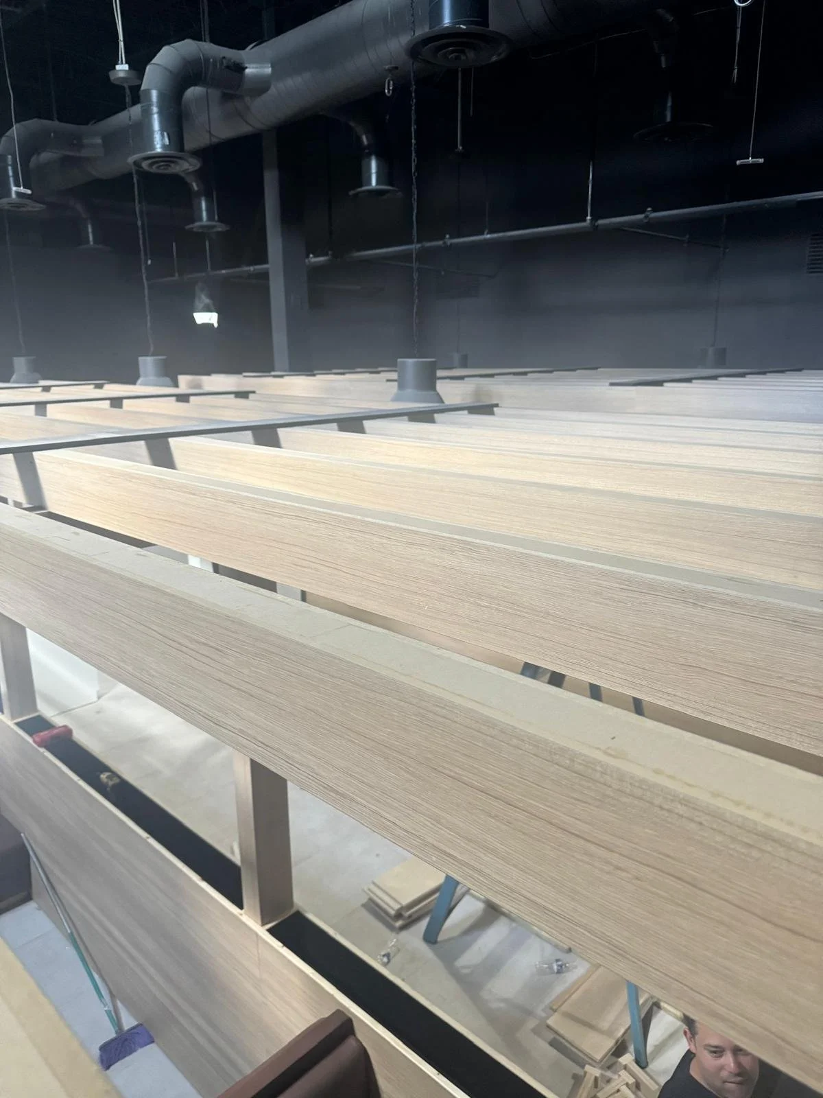 Multiple stacked wooden planks or panels in a manufacturing or storage facility with black industrial HVAC ducts and ceiling fixtures in the background.