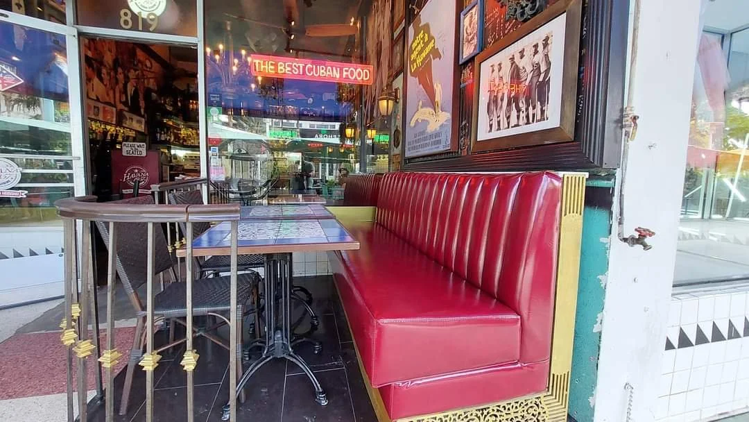 Empty indoor table with chairs next to a red upholstered booth seat at a Cuban restaurant, with posters on the wall and neon signs visible inside.