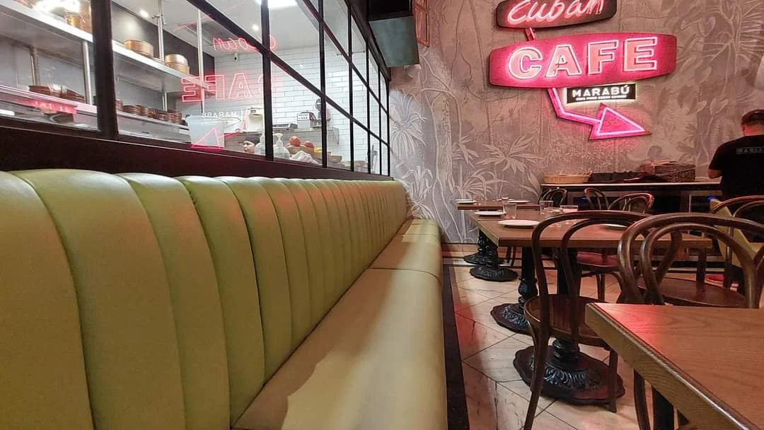 Interior of a Cuban cafe with a long yellow-green booth, wooden tables and chairs, and pink neon signs on the wall.