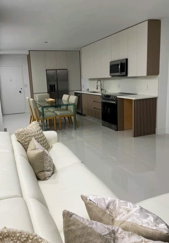 Modern open-concept kitchen and living room with white leather sofa, decorative pillows, glass dining table with beige chairs, stainless steel refrigerator, black stove, microwave, and minimalist cabinetry.