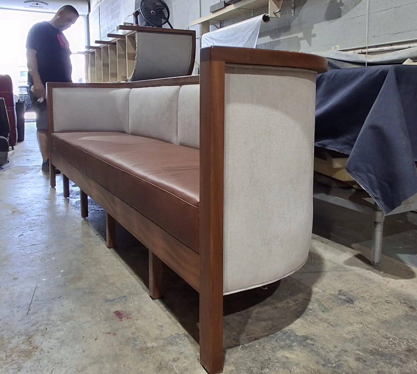A vintage-style sofa with a wood frame, beige upholstery on the sides, and brown leather on the seat cushion, placed on a concrete floor in a warehouse or workshop setting.