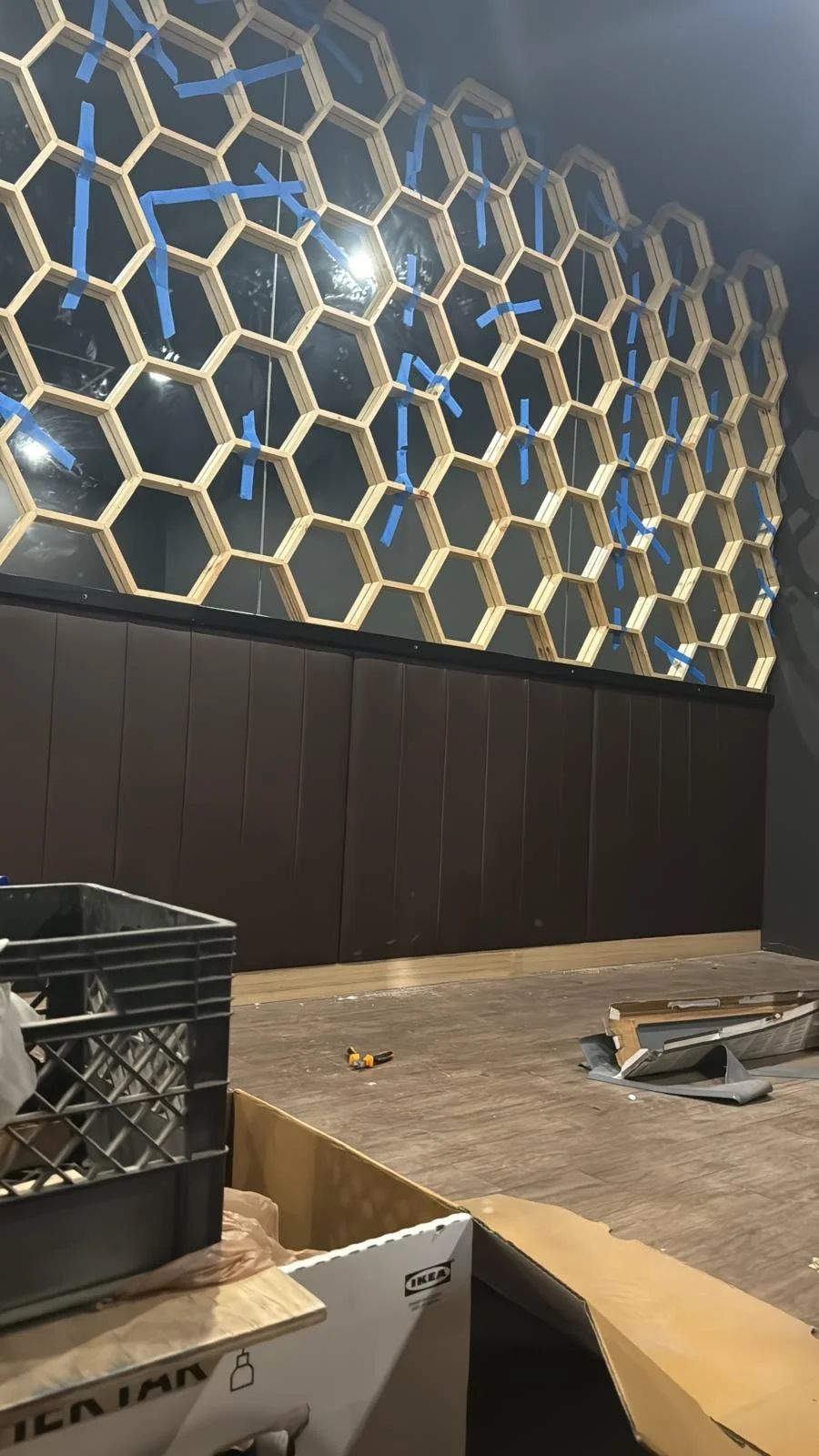 Interior of a room with a hexagonal wooden lattice wall design, blue tape markings, and construction materials on the floor.