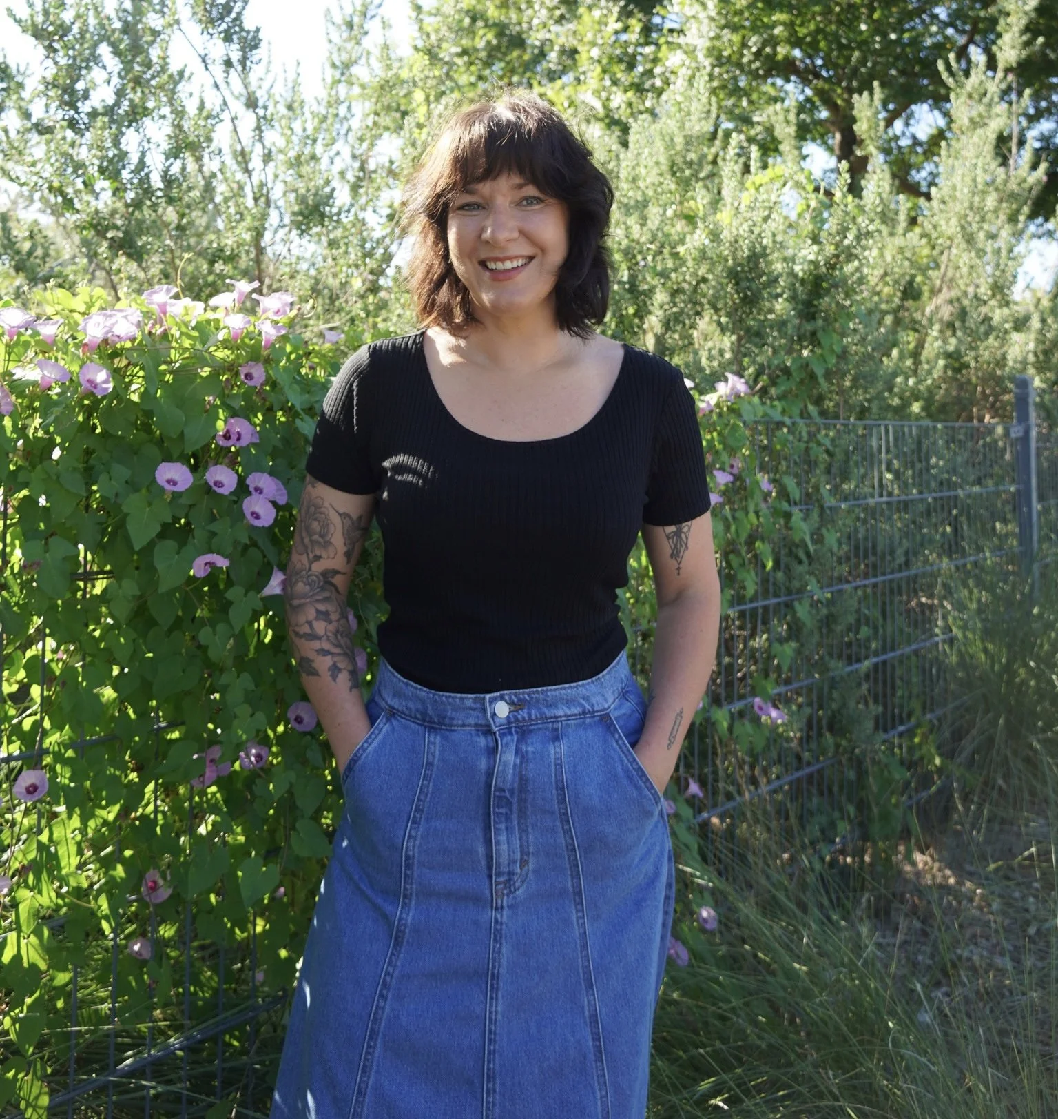 A woman with dark hair and tattoos smiling outdoors in front of greenery and pink flowers, wearing a black top and denim skirt.