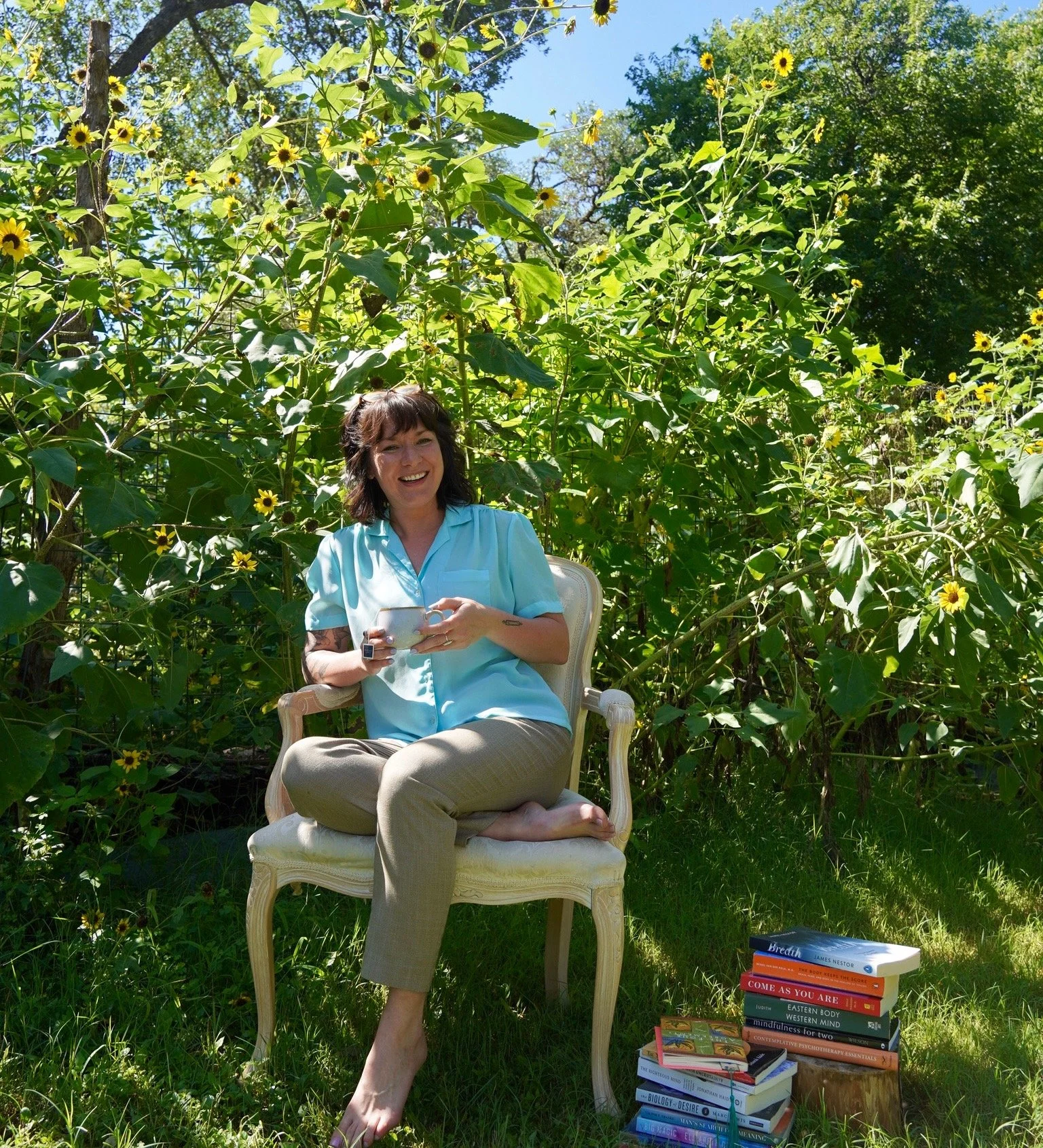 A woman sitting on a cream-colored vintage chair outdoors among lush green bushes and sunflowers, smiling and holding a mug, with stacks of books on the ground nearby.