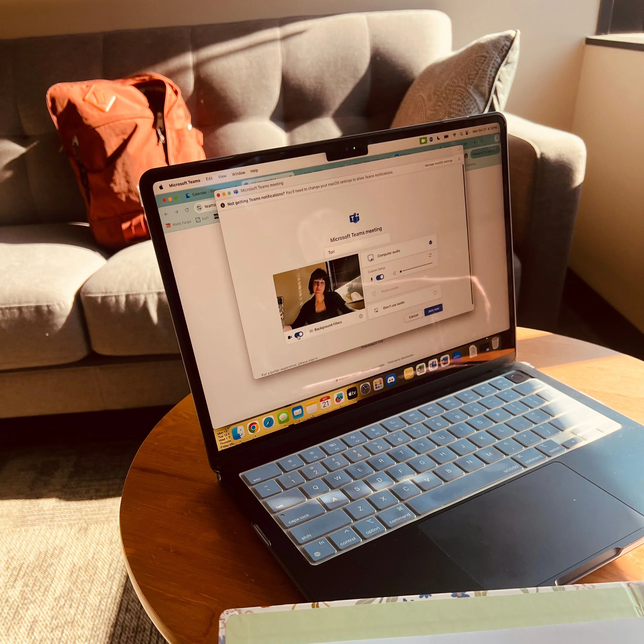 Open laptop on a wooden table displaying a Microsoft Teams meeting screen. A red backpack is on the sofa in the background, which has a beige cushion. Sunlight is casting shadows in a cozy living room.