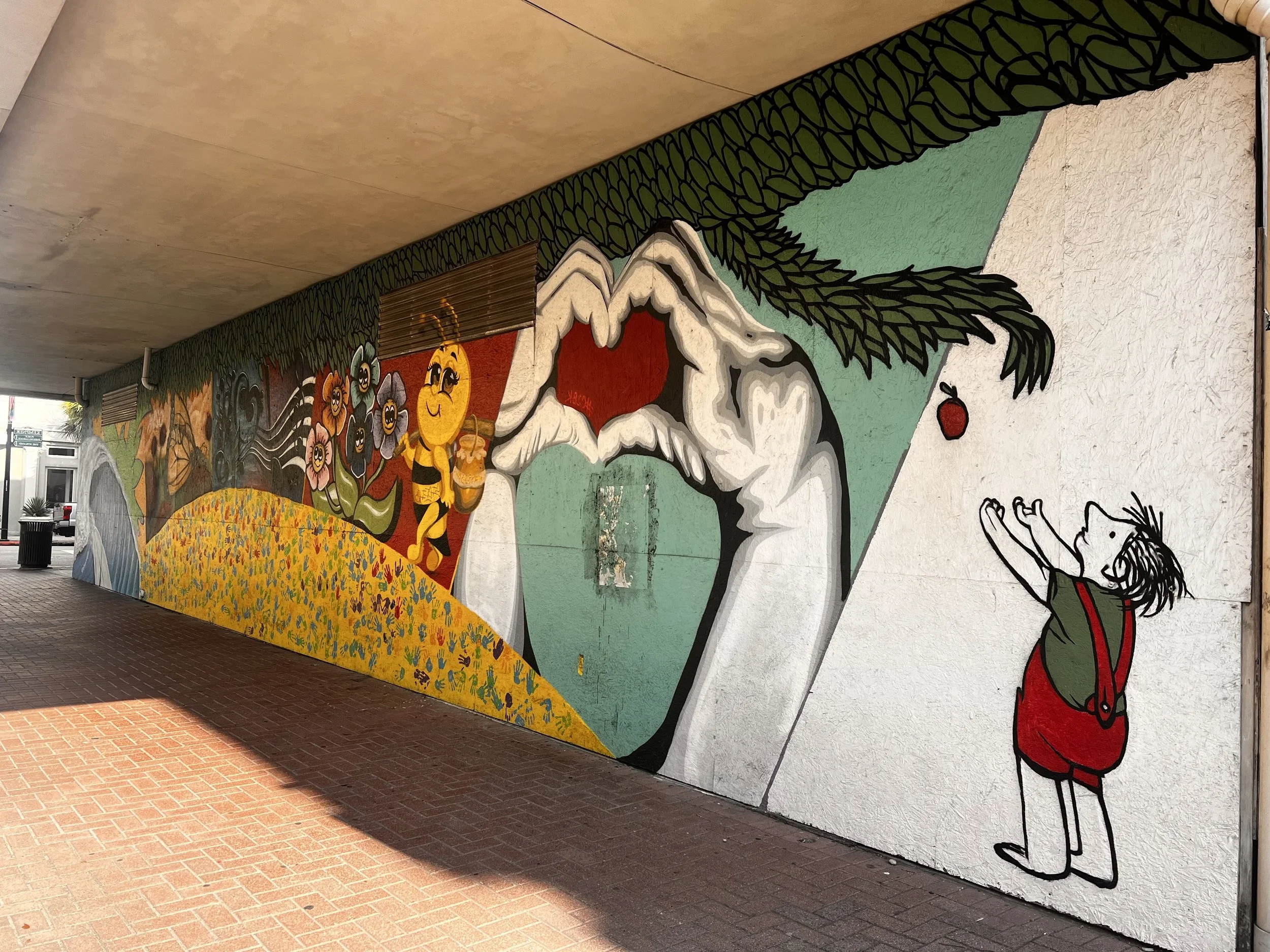 Colorful mural on a wall depicting a cartoon scene with a woman on the ground trying to catch a falling apple, a large eye with a hand forming a frame, bees, flowers, and a small figure with outstretched arms reaching towards the apple.