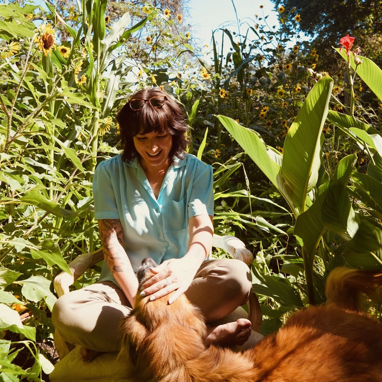 A woman sitting cross-legged in a lush garden with tall green plants and yellow flowers, smiling and petting a large, fluffy dog lying on the ground.