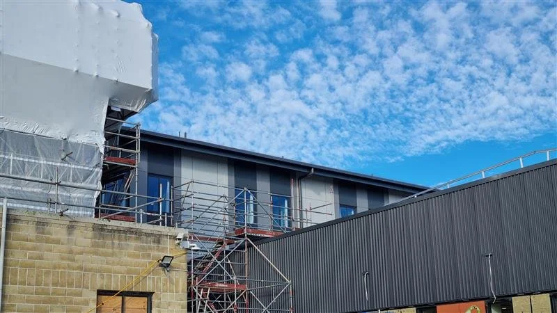 Construction site with scaffolding around a modern building, blue sky with scattered clouds in the background.