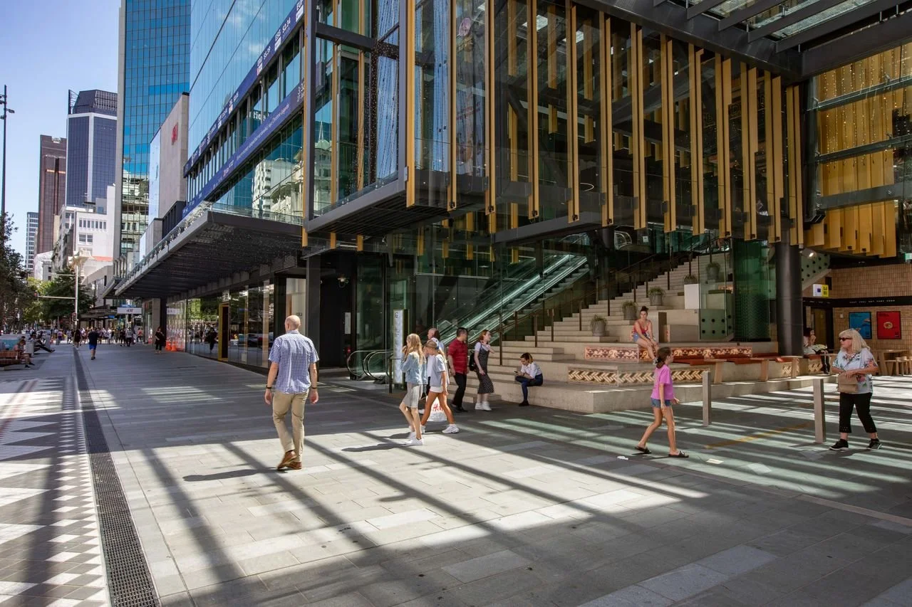 City street scene with pedestrians crossing at a crosswalk near a modern glass building with yellow accents and an outdoor staircase.