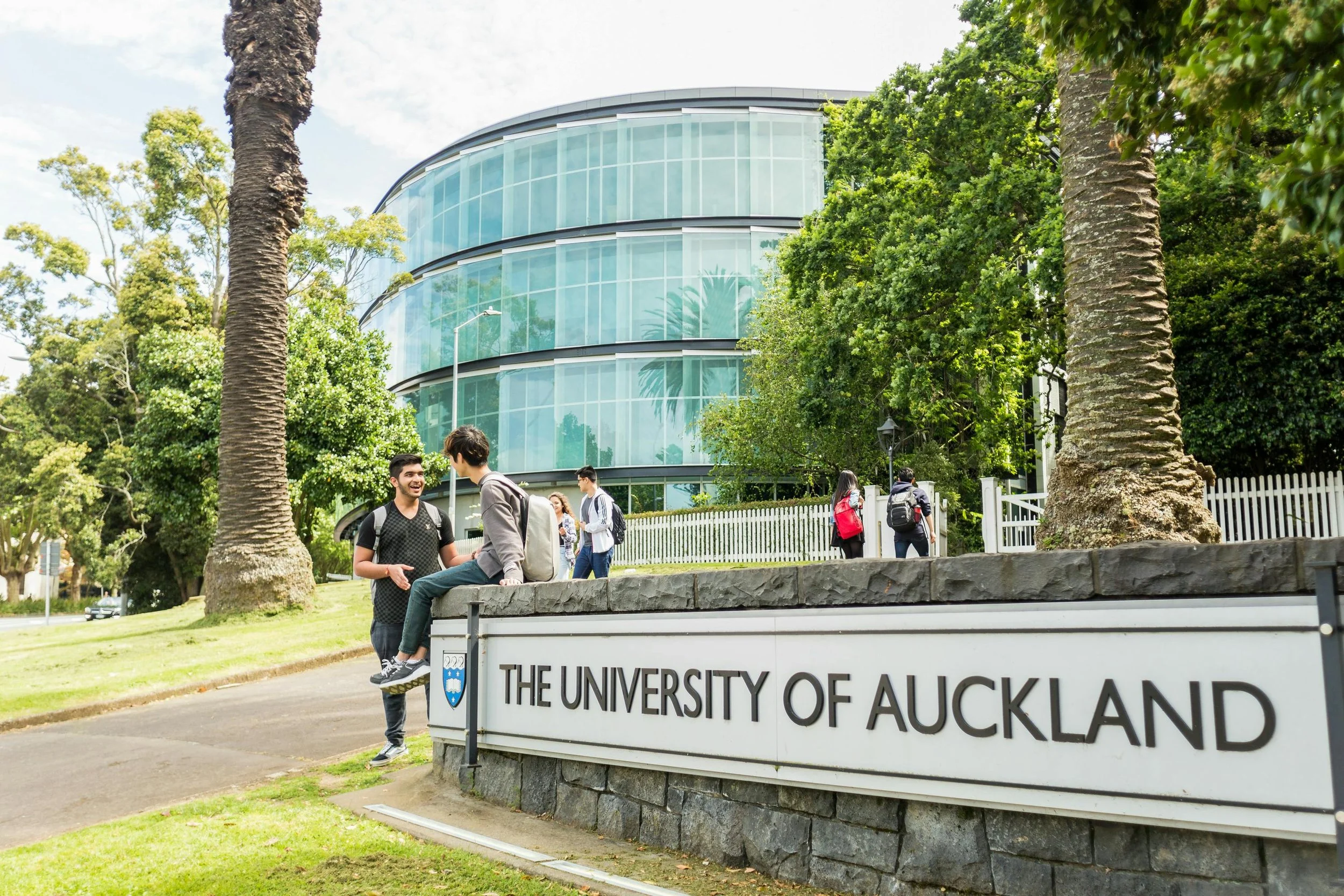 Students on campus near a sign that reads "The University of Auckland" in front of a modern glass building with trees and greenery around.