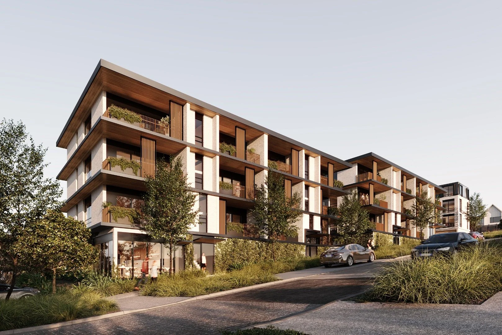 Modern apartment complex with multiple stories, balconies, and greenery, situated along a sloped road with cars parked and trees in the landscape.