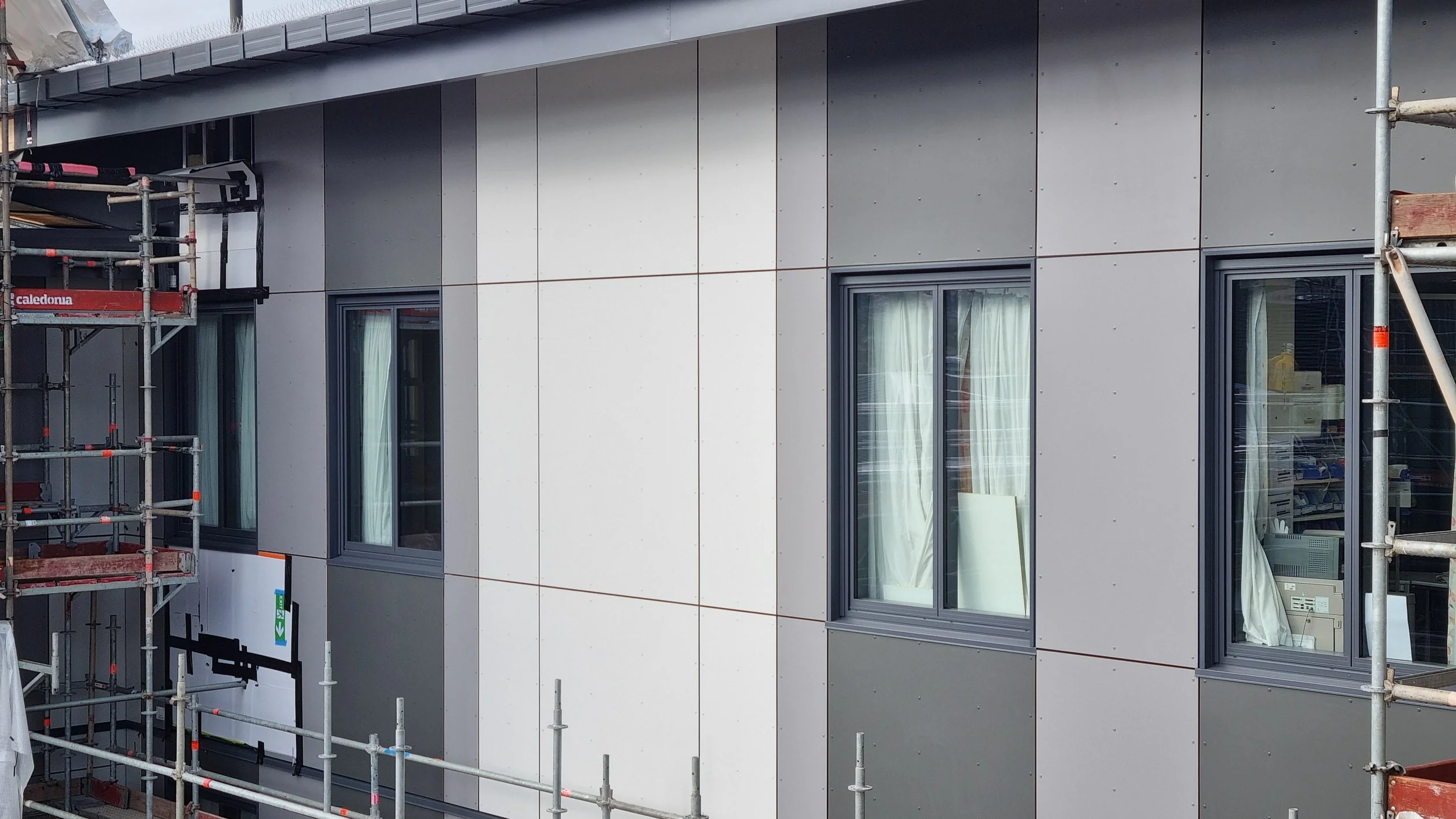 Building exterior under construction with installed gray and white panels and visible windows, scaffolding on the sides.