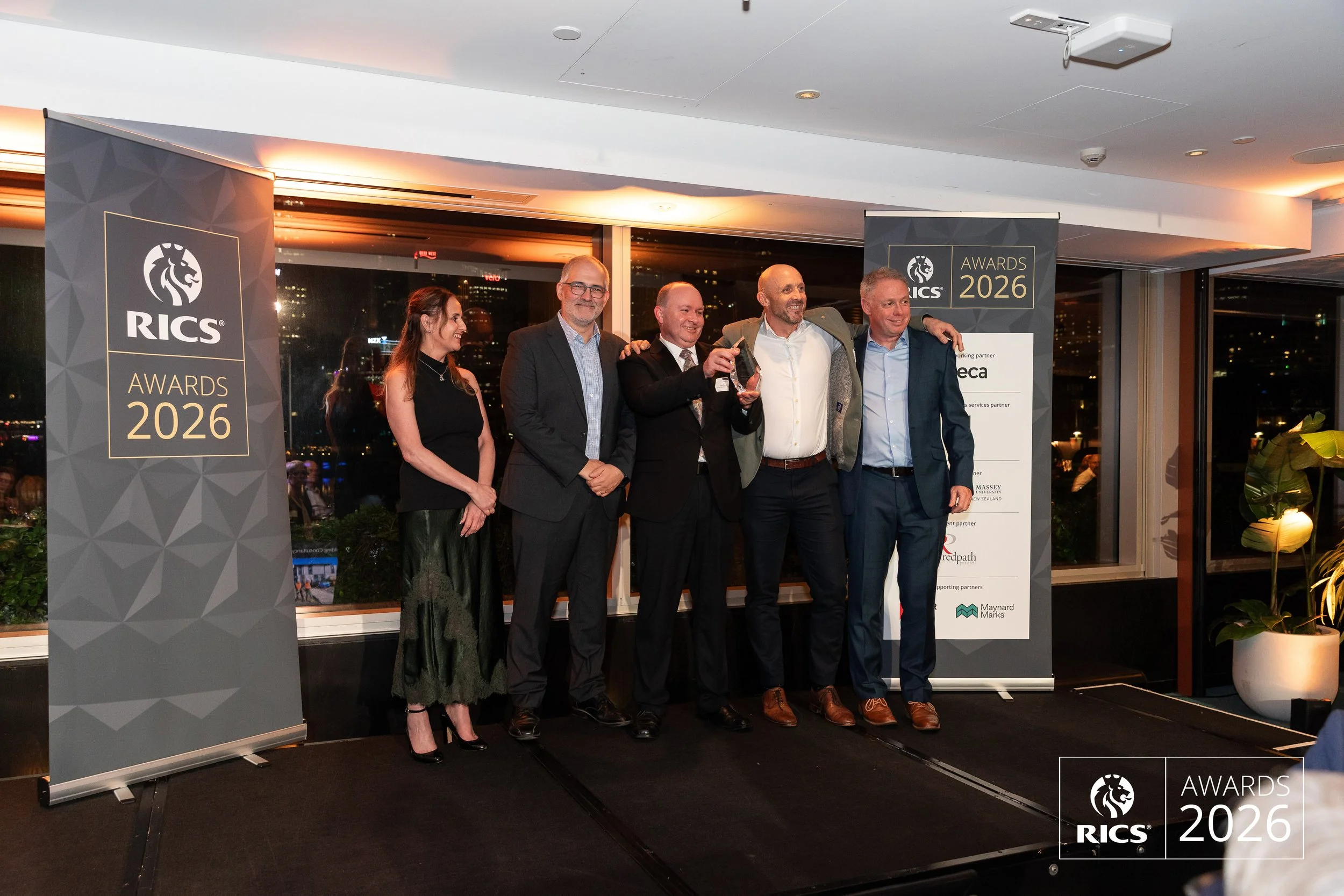 Group of five people standing together at the RICS Awards 2026, some holding an award, with a city view through large windows behind them.