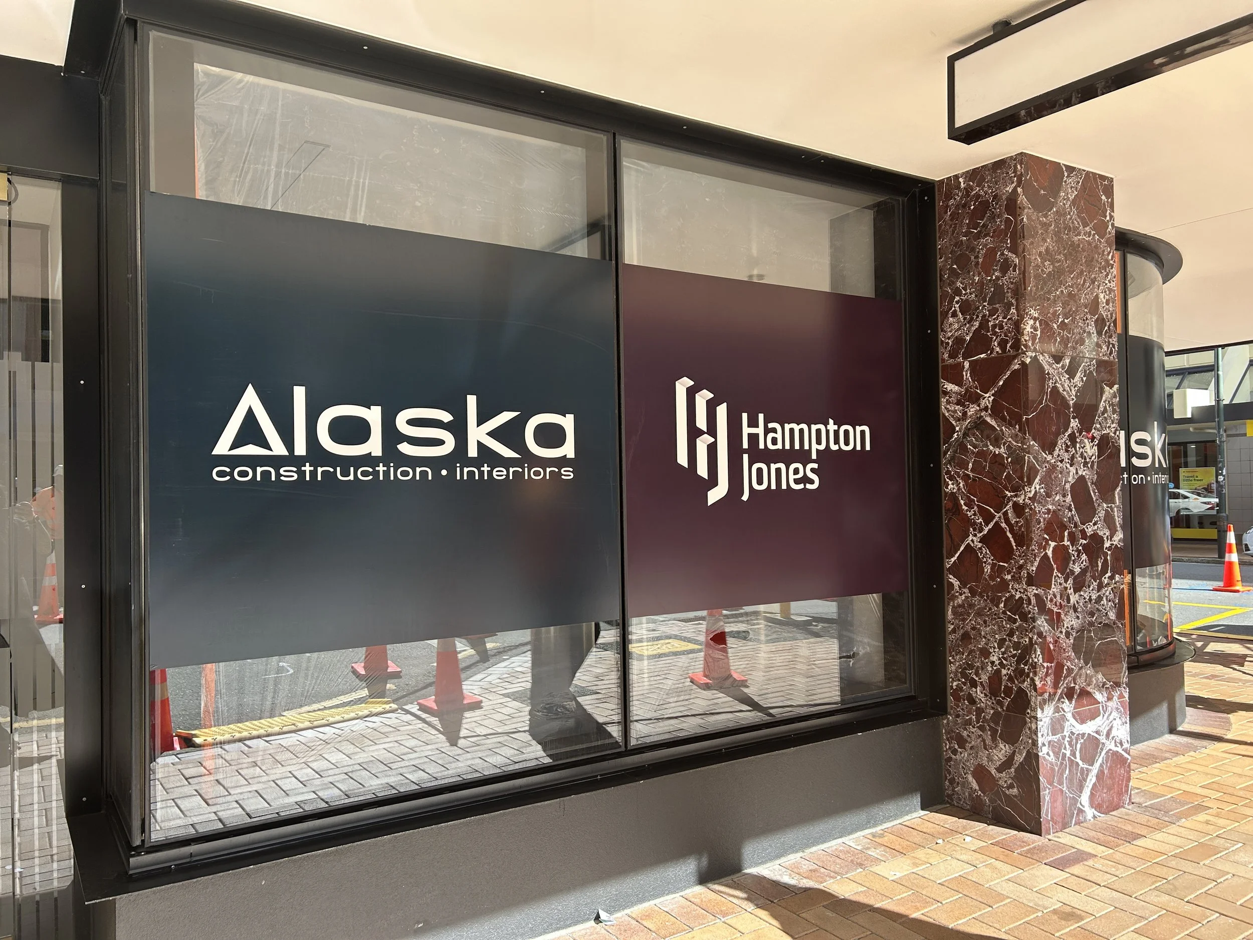 Storefront windows with business signs for Alaska Construction Interiors and Hampton Jones, featuring a marble column and brick sidewalk outside.