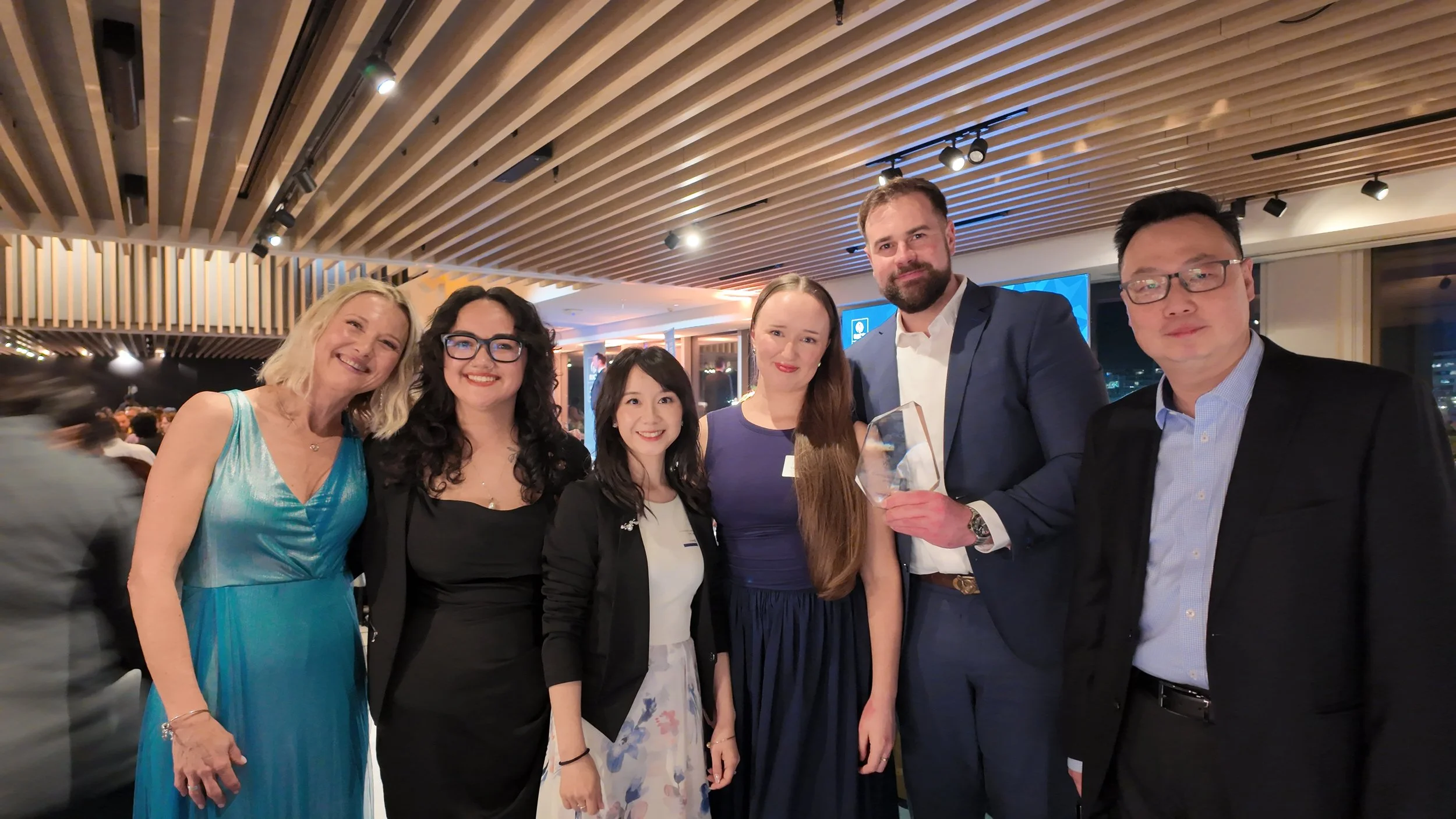 Hampton jones quantity surveyors standing together at RICS awards event, smiling at the camera, with a wooden ceiling and screens in the background.