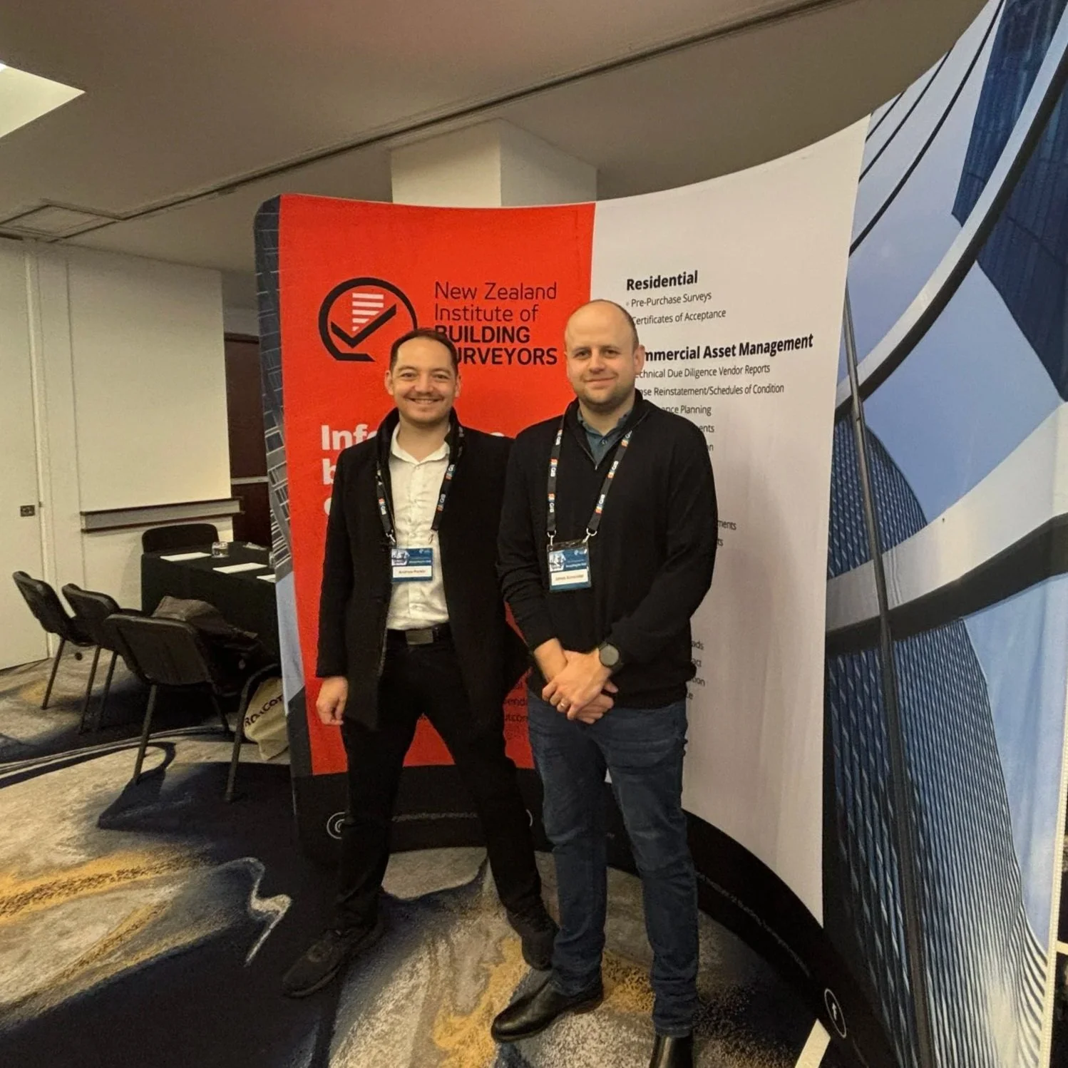 Two hampton jones building surveyors standing indoors in front of a backdrop with the logo and information of the New Zealand Institute of Building Surveyors, smiling and wearing conference badges.