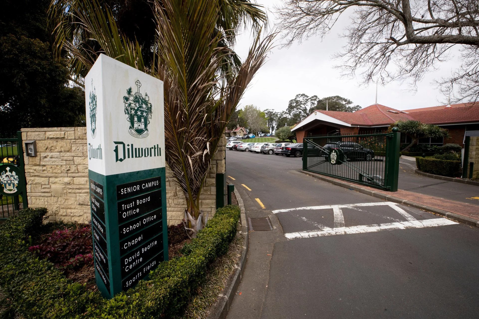 Sign at Dilworth School campus with various administrative offices and sports fields, with cars parked in the background and an open gate leading to the campus.