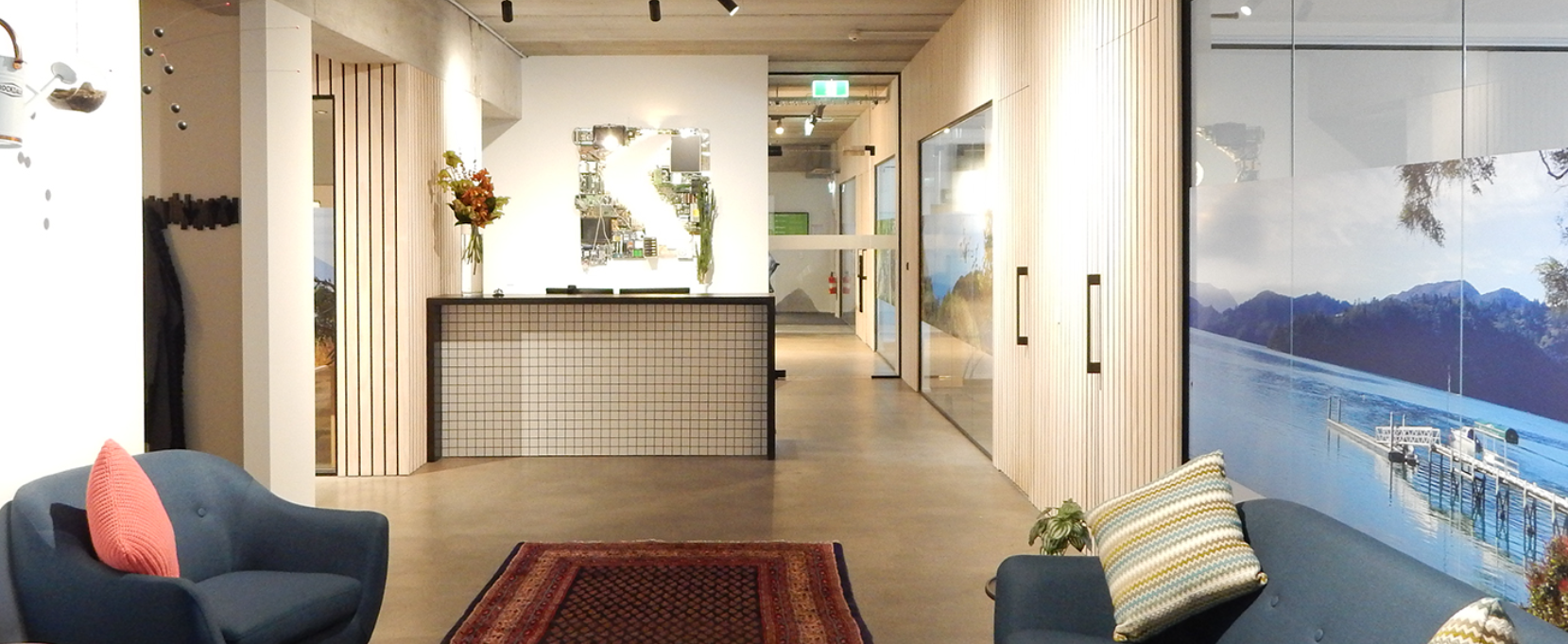 Modern lobby with blue sofas, colorful pillows, a patterned rug, a reception desk, and large wall mural of a lake with mountain backdrop.