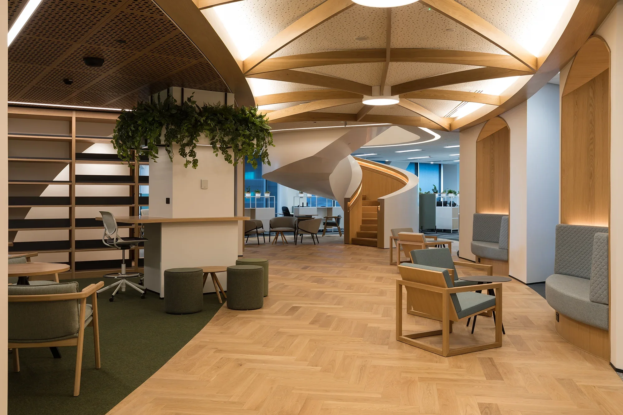 Modern office lounge area with wooden flooring, several chairs, a spiral staircase, plants, and curved architectural design elements.
