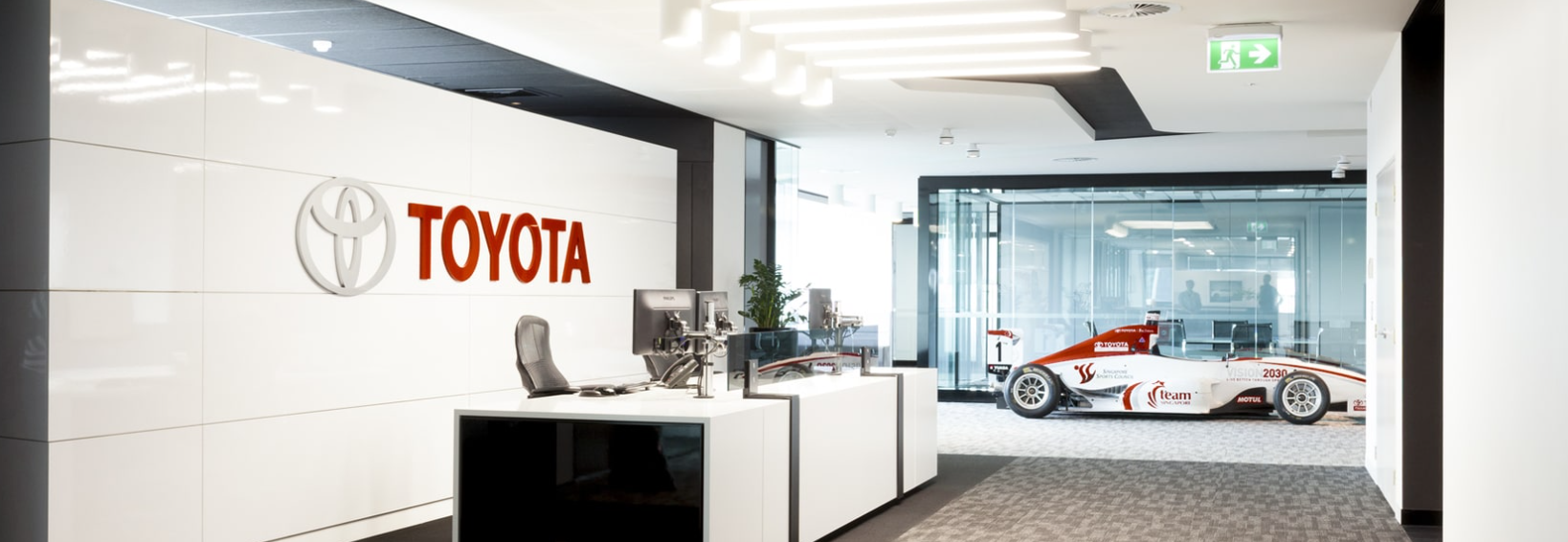 Toyota office with a race car displayed behind glass, a reception desk with two computer monitors, and a white wall with the Toyota logo in the foreground.
