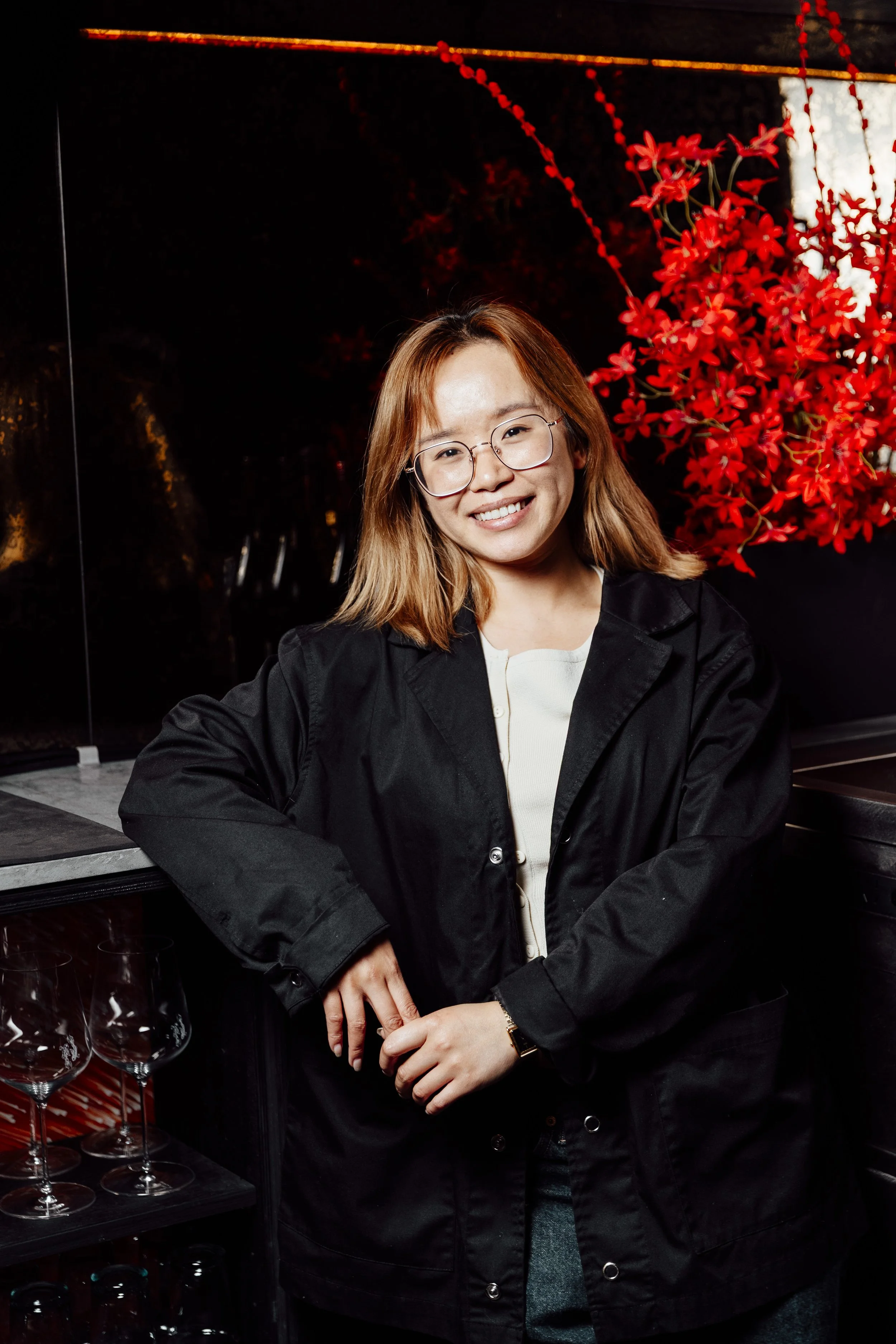 A woman with glasses and shoulder-length hair smiling, leaning against a bar with empty wine glasses, with a red floral decoration in the background.