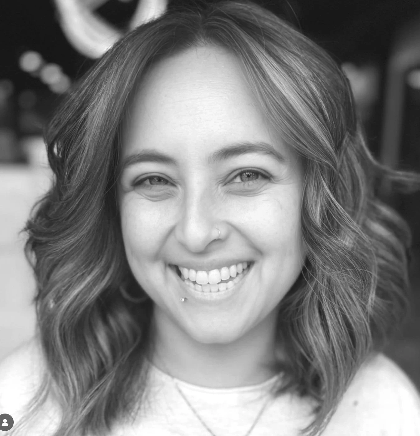 A smiling woman with wavy hair, a nose piercing, and a labret piercing, looking directly at the camera. Black and white photo.