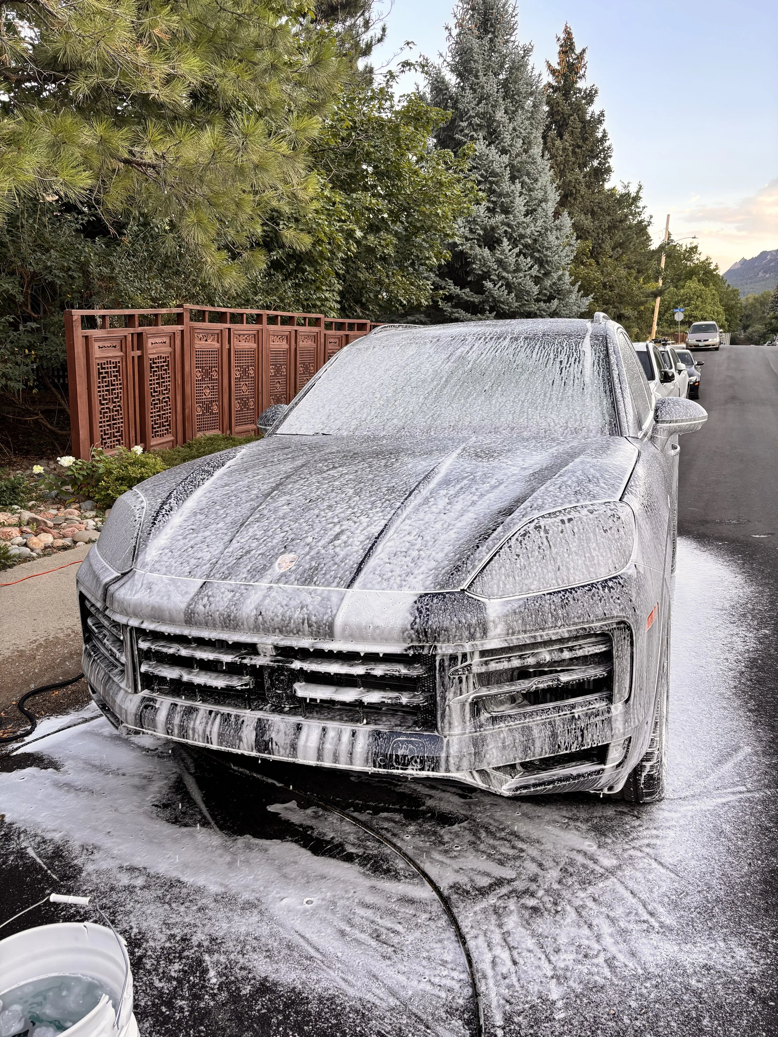 A silver Porsche SUV is being washed and covered in soap suds, with a bucket nearby, on a driveway with trees and a red fence in the background.