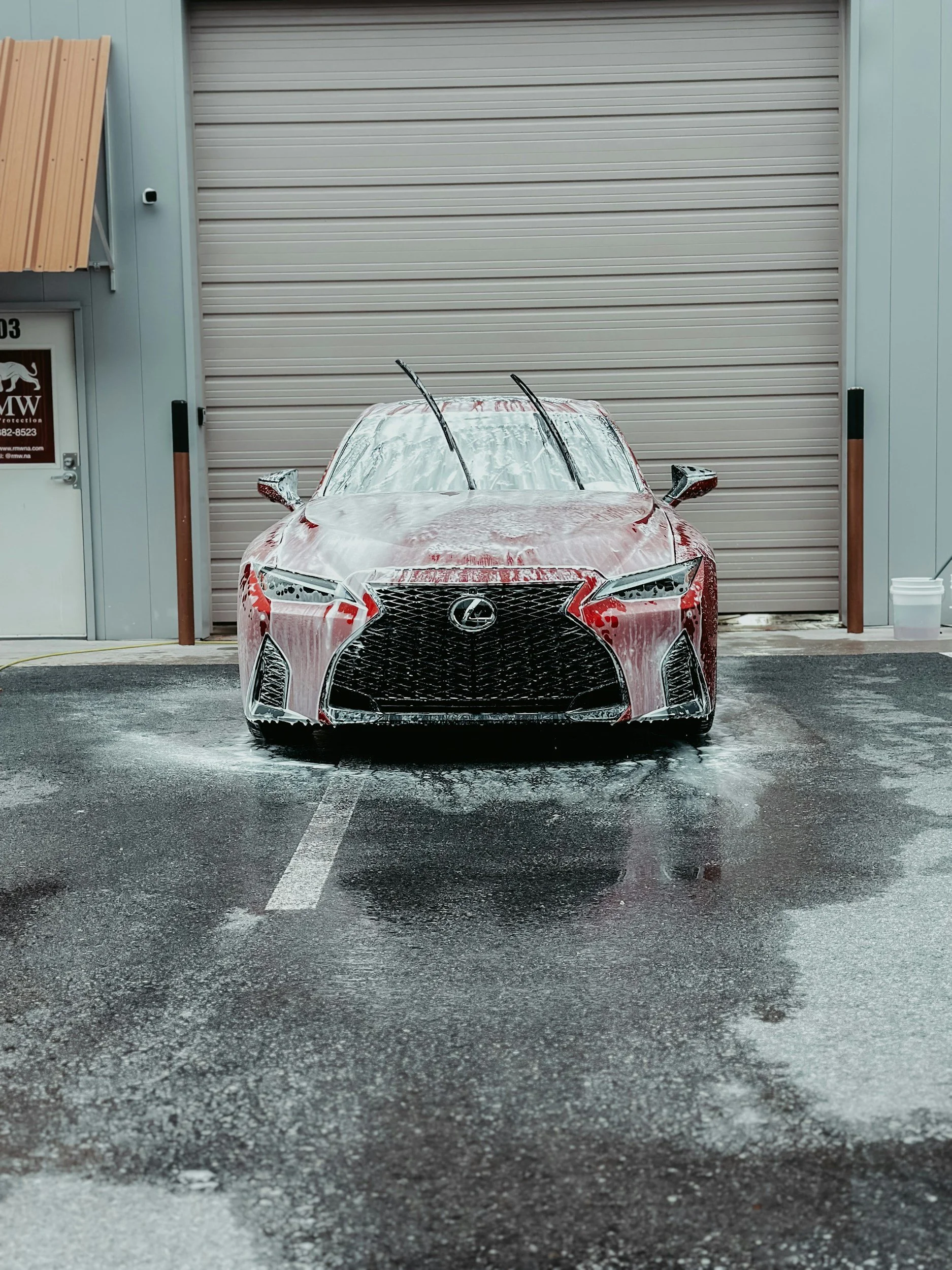 Red Lexus sports car being washed in an outdoor car wash station with soap and water, parked in front of a closed garage door.