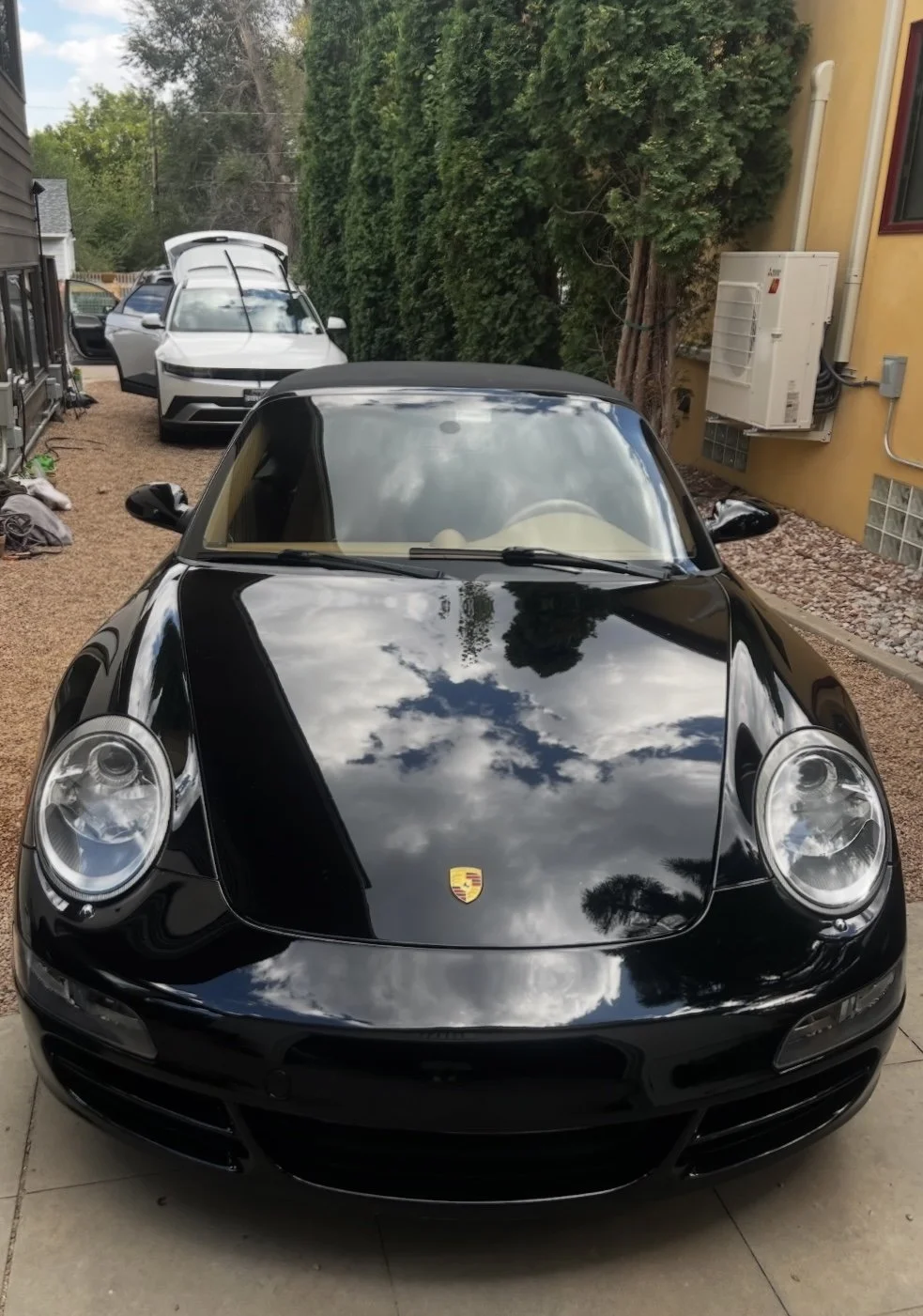 A black Porsche sports car with a reflected sky on its hood, parked in a driveway in front of a yellow house, with a white SUV and another car in the background.