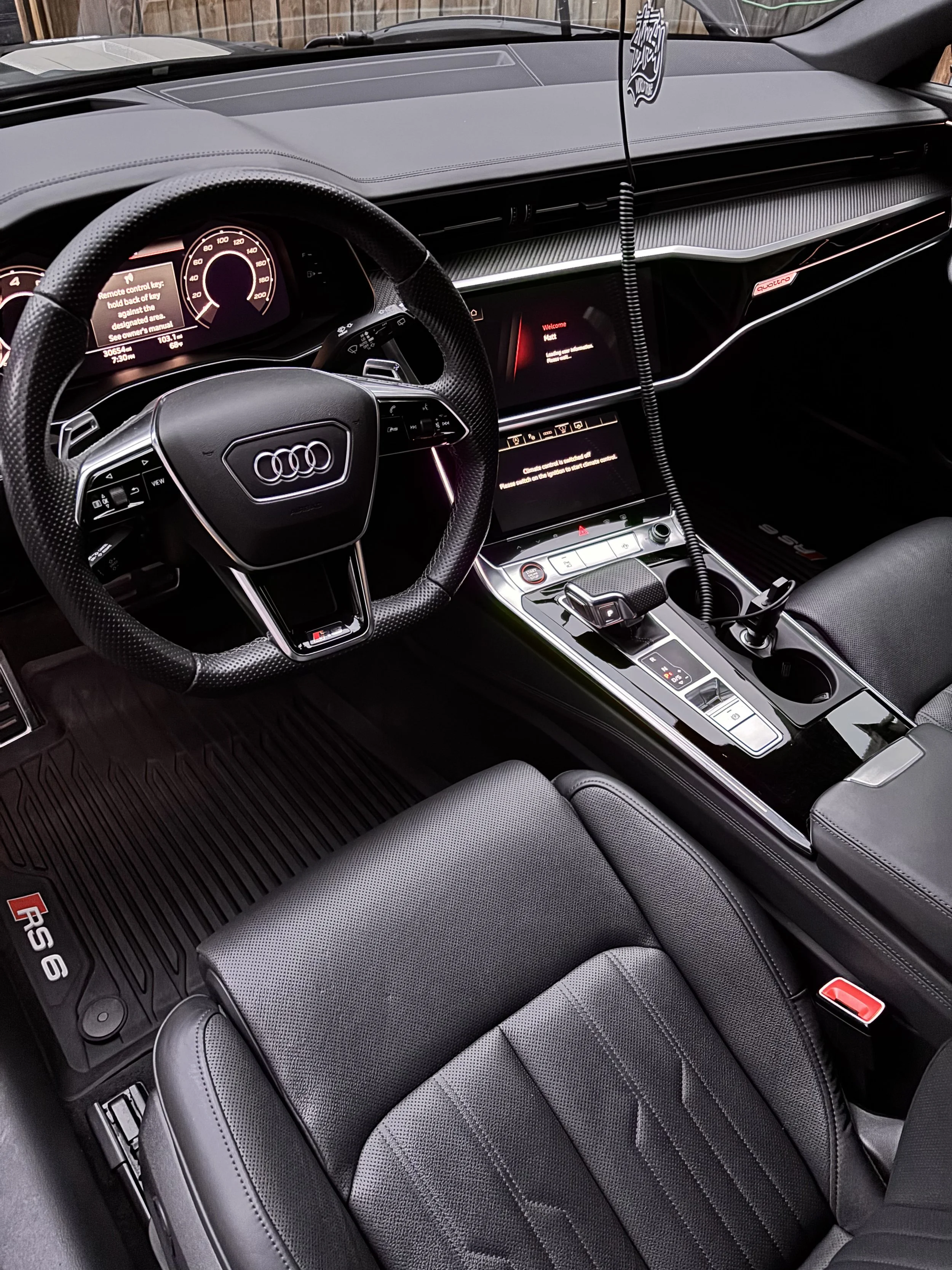 Inside view of an Audi RS6 interior featuring a steering wheel with the Audi logo, digital instrument cluster, central infotainment screen, gear shift, and control buttons, with black leather seats and floor mats.