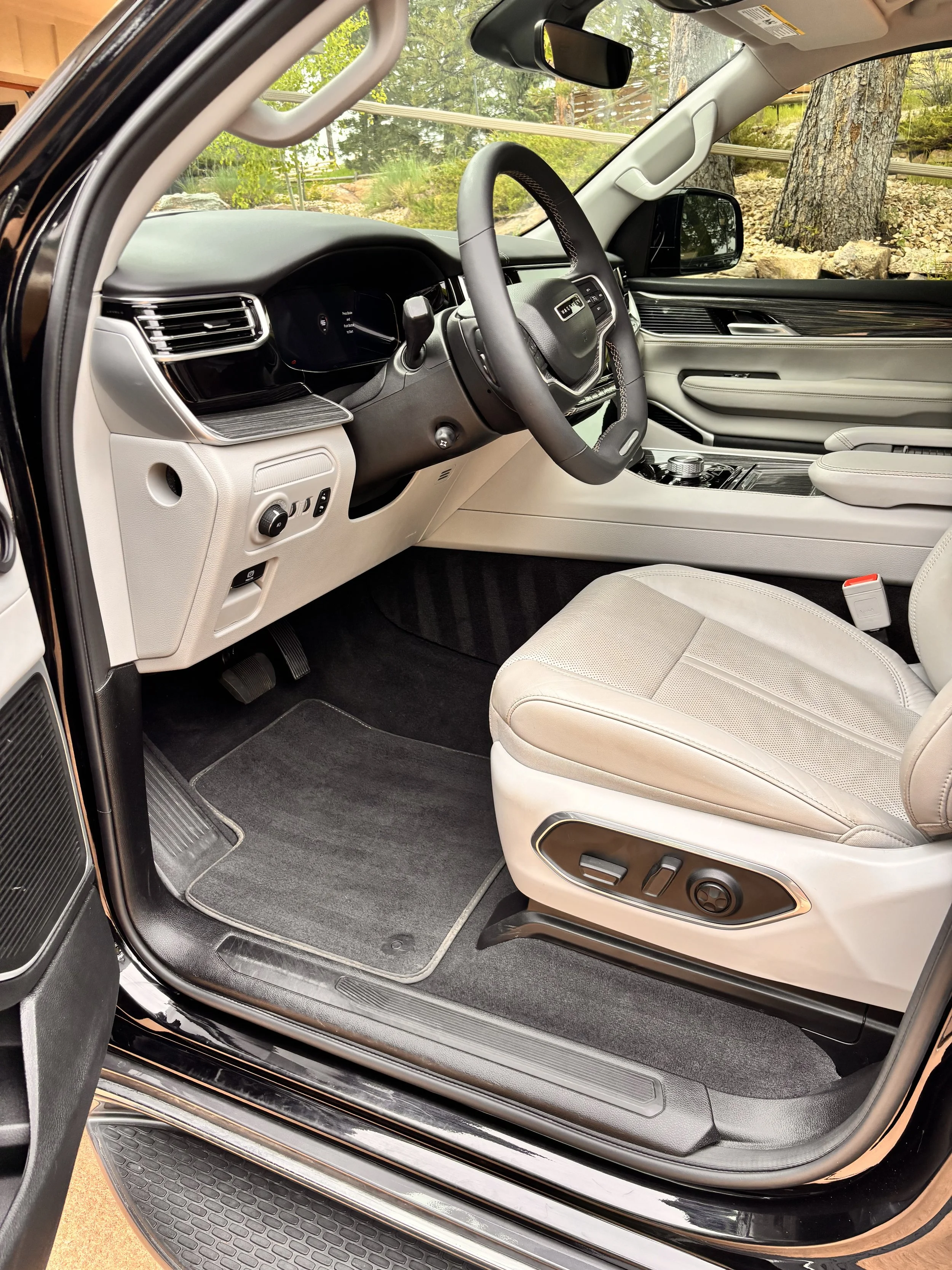 Interior of a luxury vehicle's front passenger area with beige seats, a black dashboard, and a steering wheel, parked outdoors with trees visible through the windshield.