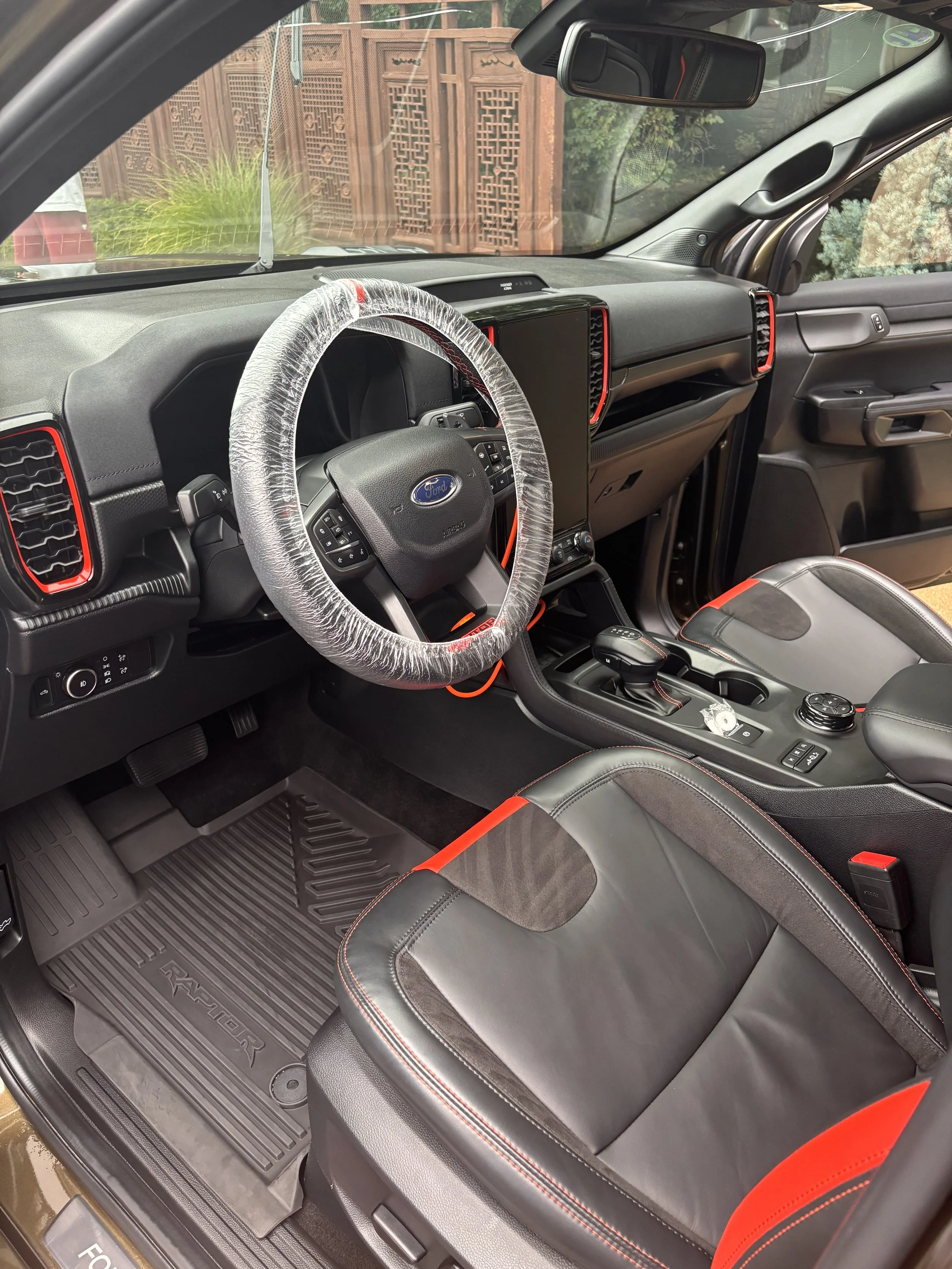 Interior of a Ford vehicle, showing the dashboard, steering wheel with a plastic cover, gear shift, and black and red leather seats.