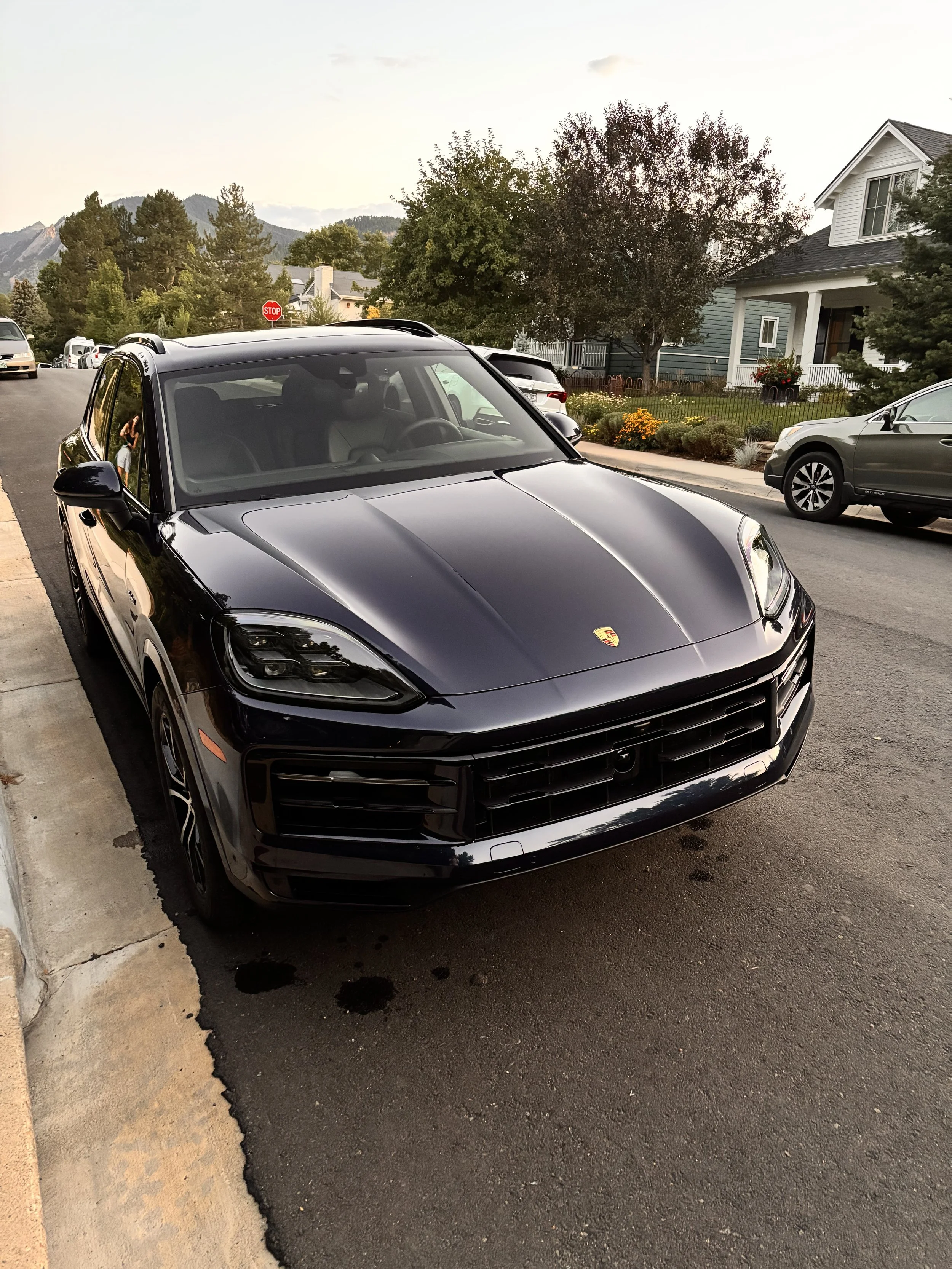 Black Porsche SUV parked on the side of a residential street with houses, trees, and mountains in the background during daytime.