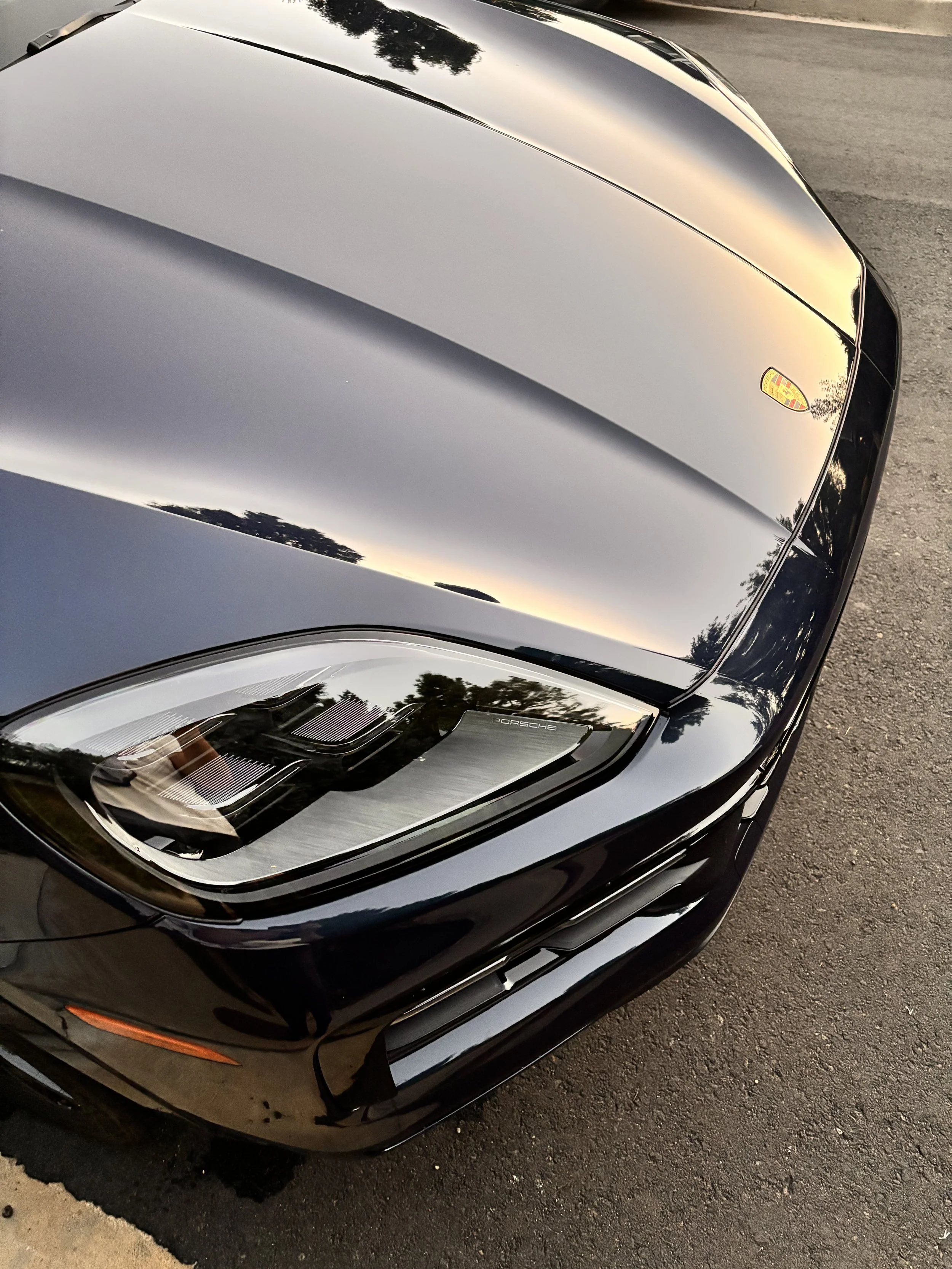 Close-up of a black Porsche sports car's front end, showing the hood, headlight, and part of the front bumper, with reflections of trees and the sky on the surface.