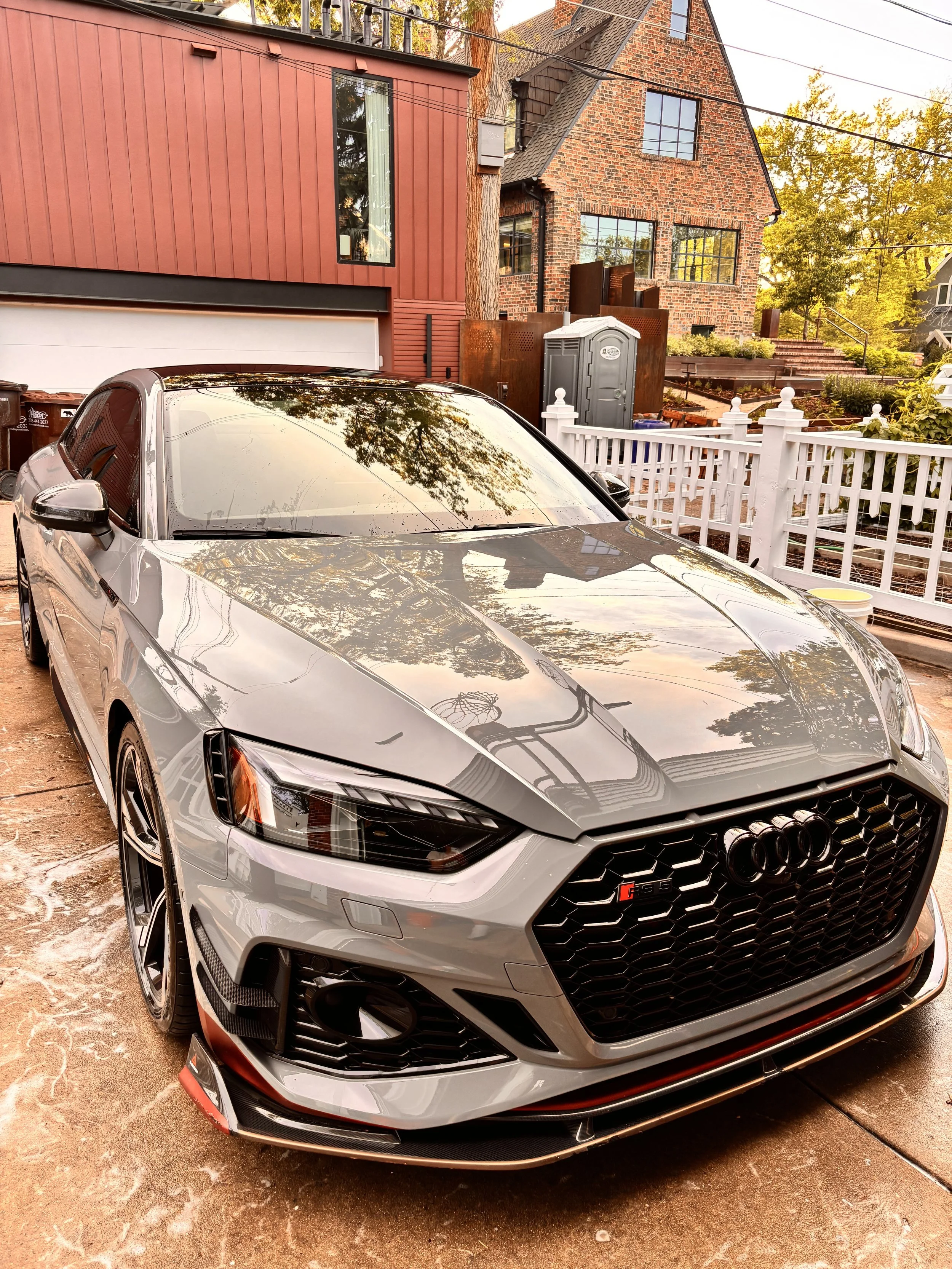 A silver Audi RS5 car parked on a driveway in front of a house with a red wooden exterior on the left and a brick house with large windows in the background. The driveway has some soap suds, indicating washing or cleaning.