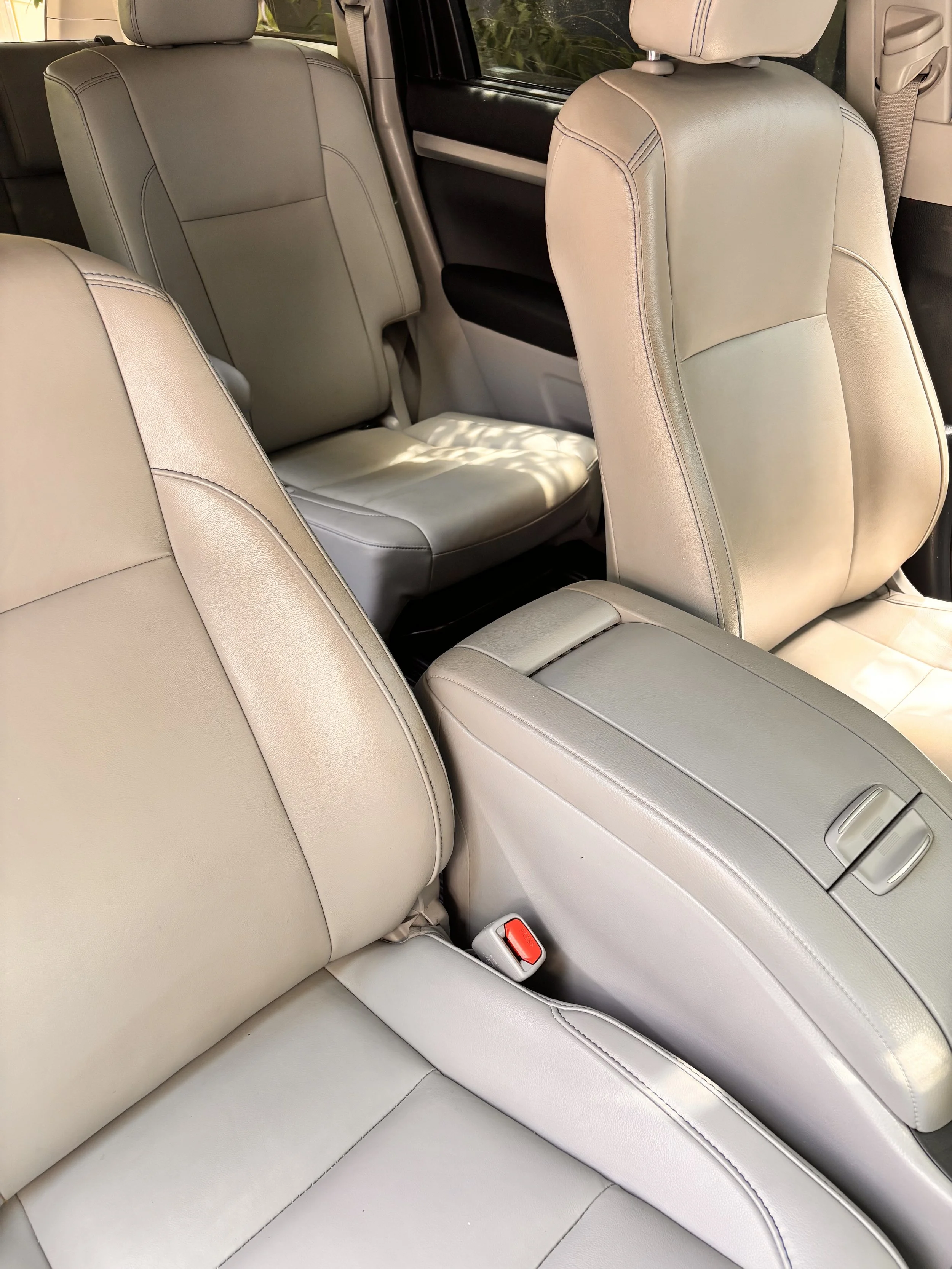 Interior of a vehicle showing beige leather seats, including the front passenger seat, the middle row, and the back row, with sunlight casting shadows inside.
