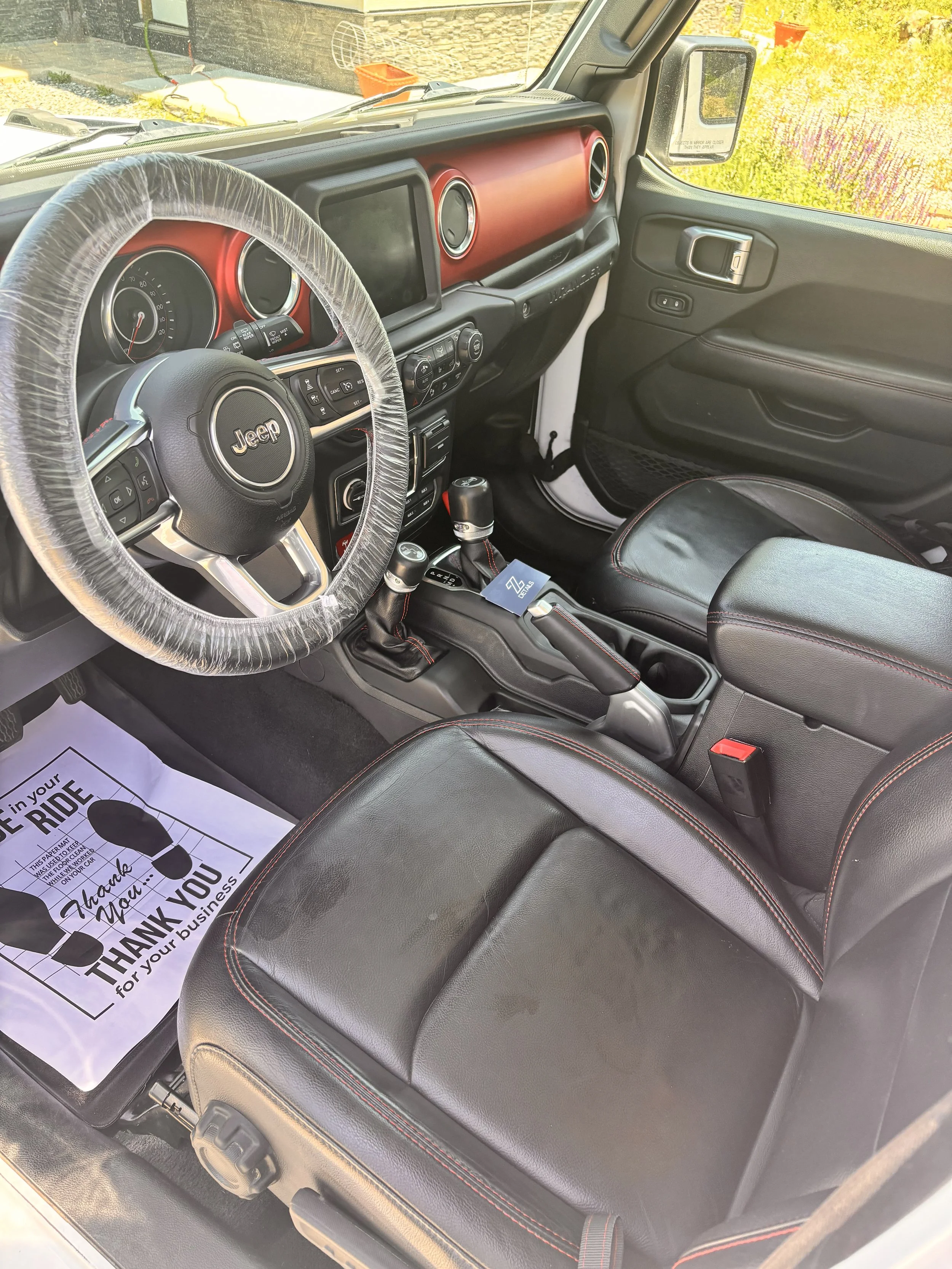 The interior of a Jeep vehicle with a black and red dashboard, black leather seats with red stitching, and a steering wheel wrapped in protective plastic. There is a white paper mat on the driver's side with a thank you message for the business.