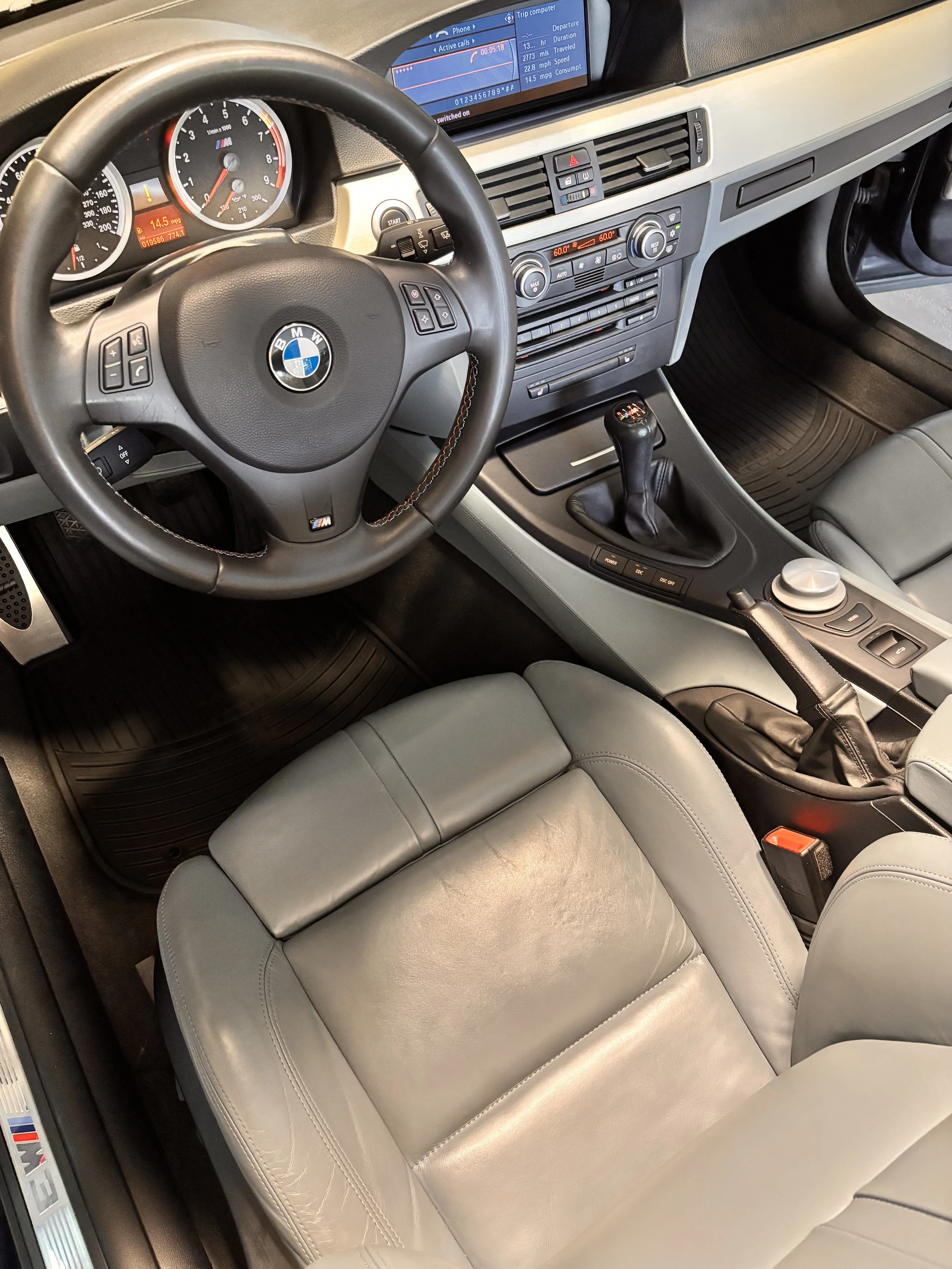 Interior of a BMW car, showing the steering wheel, dashboard, gear shift, and front seat with gray leather upholstery.