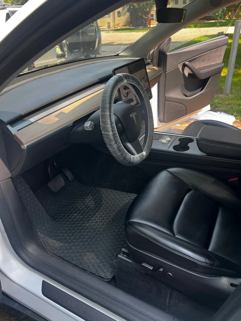 Interior of a Tesla vehicle, showing the dashboard, steering wheel wrapped in plastic, black leather seat, and a chamois-lined floor mat.