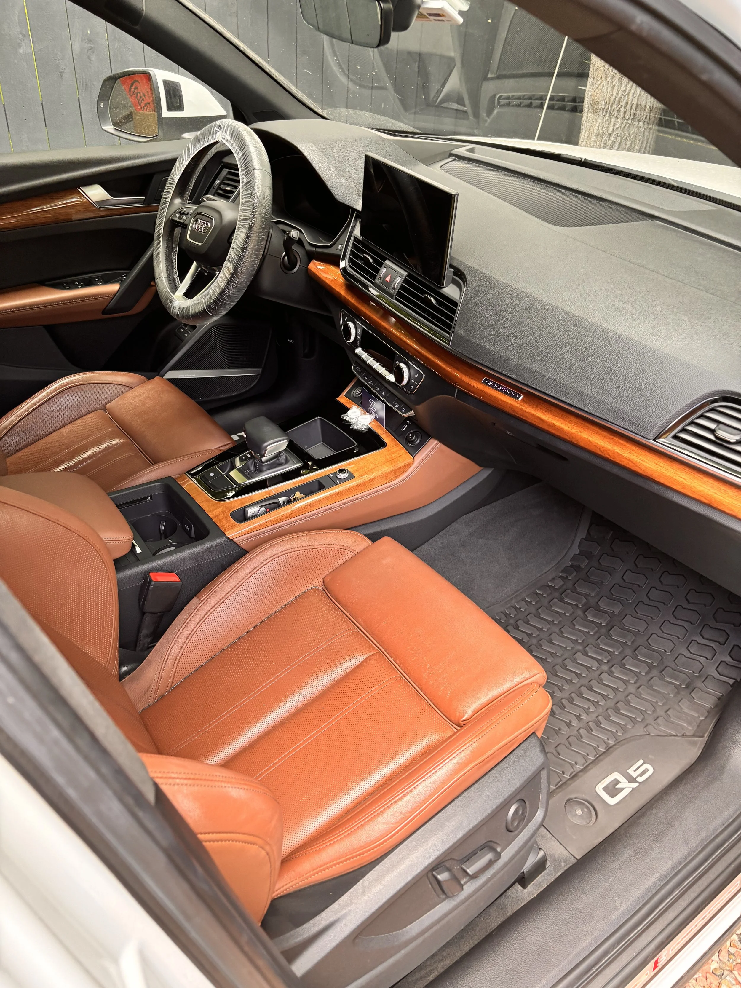 Interior of a luxury car with leather seats, a steering wheel with an Audi logo, wooden trim on the dashboard and center console, digital display screen, gear shifter, and floor mat with 'Q5' branding.