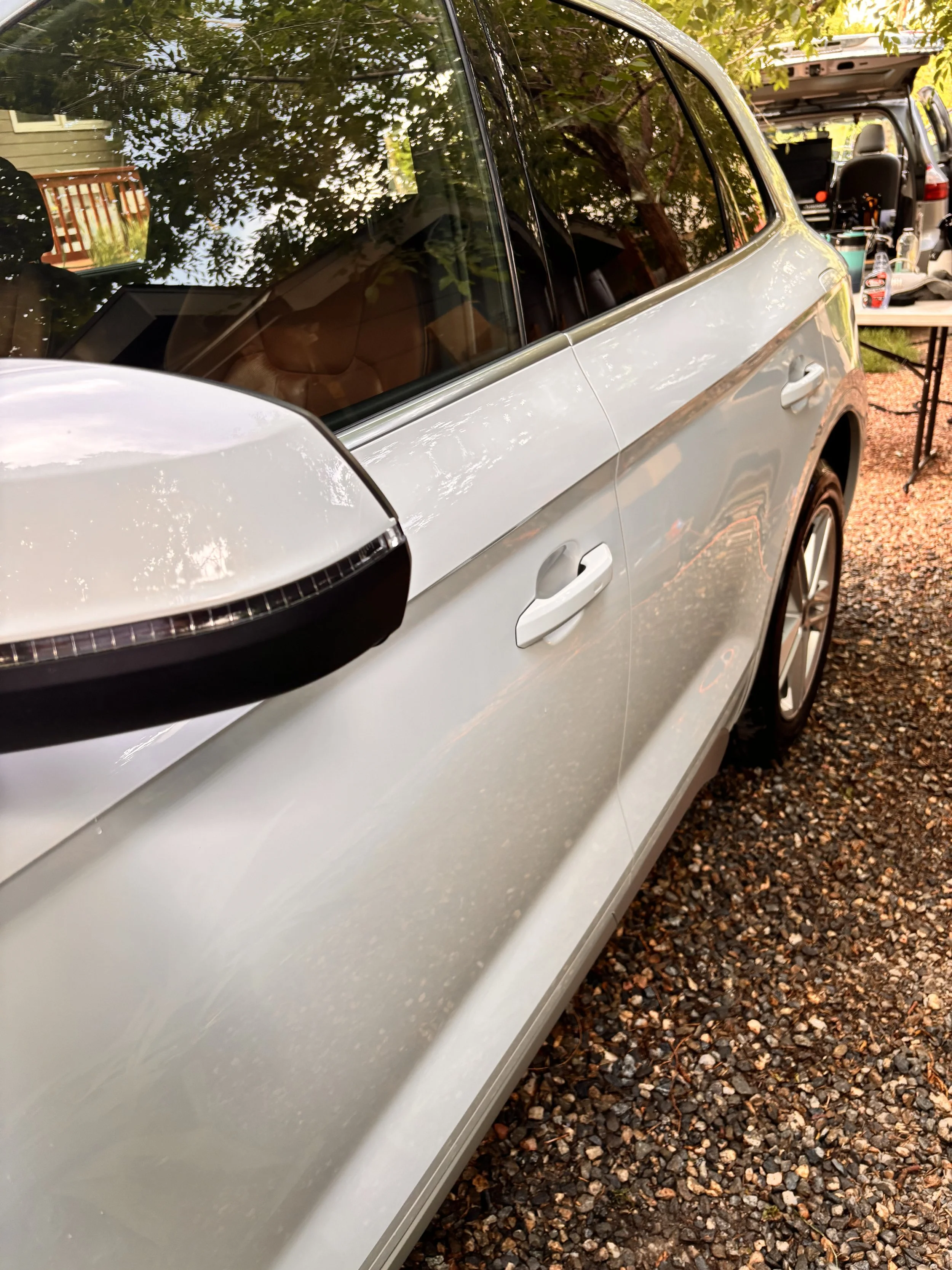 Close-up of a silver car parked on a gravel surface, showing the side mirror, windows, and part of the door.