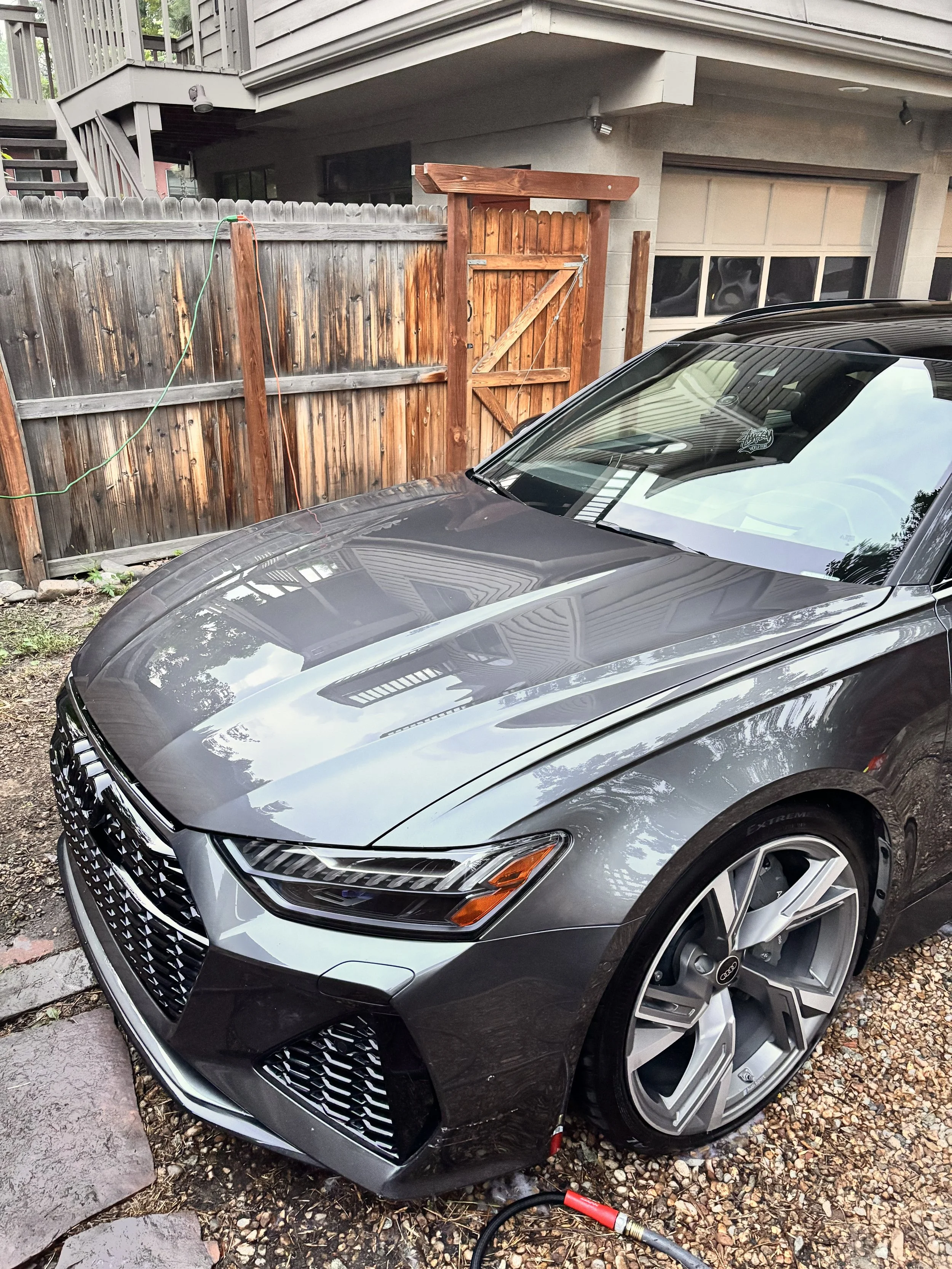 A gray Audi car parked in a residential driveway with a wooden fence and house in the background.