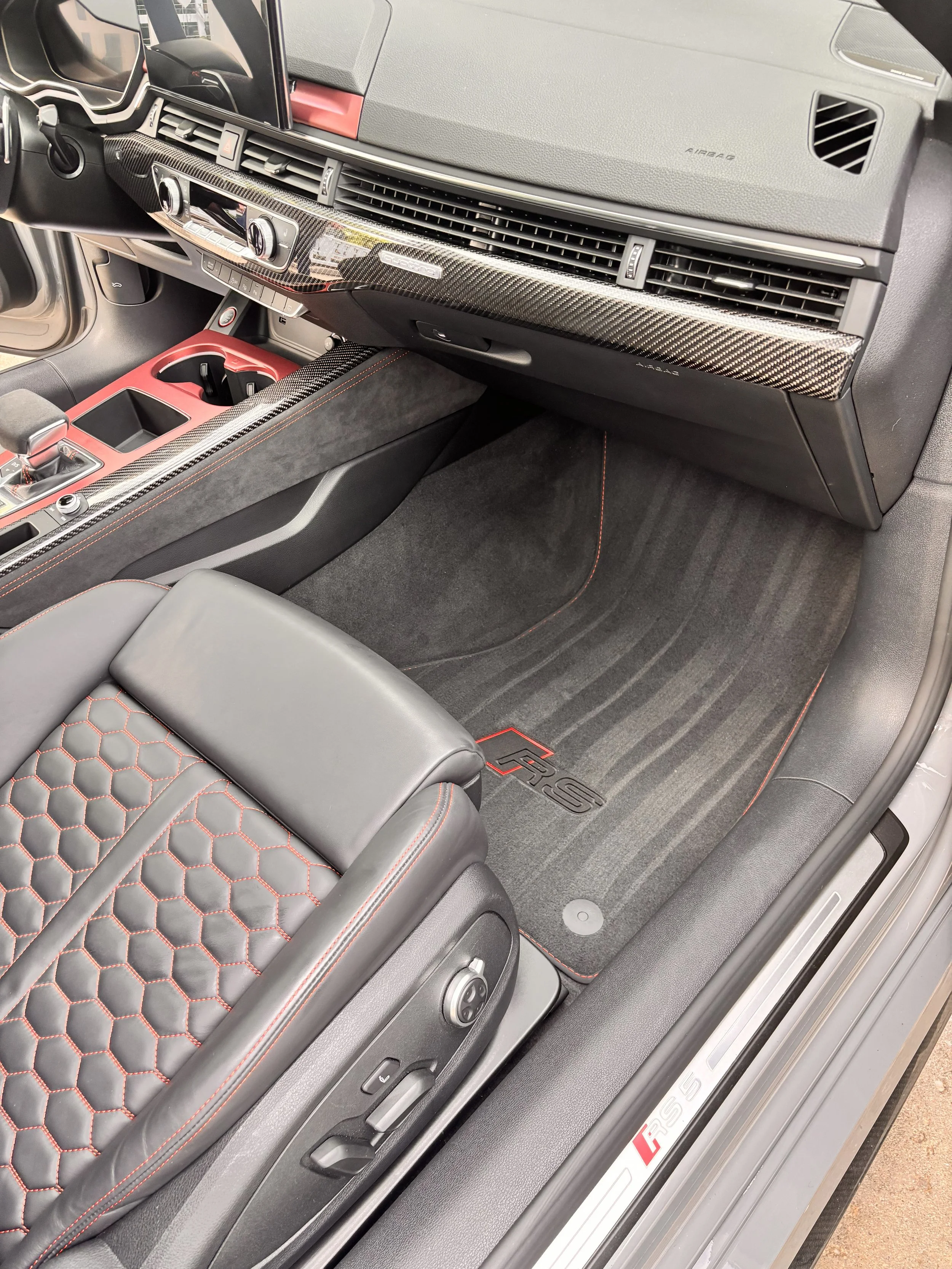 Interior of a car showing the front passenger side with a black floor mat, a partially visible seat with honeycomb stitching, and a dashboard with a carbon fiber panel.