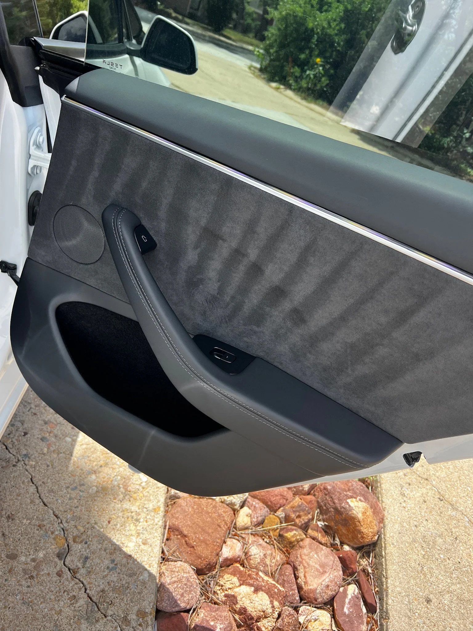 Close-up of a car door interior with black and gray design, including speaker, window control, and handle, with pavement and rocks outside the vehicle.