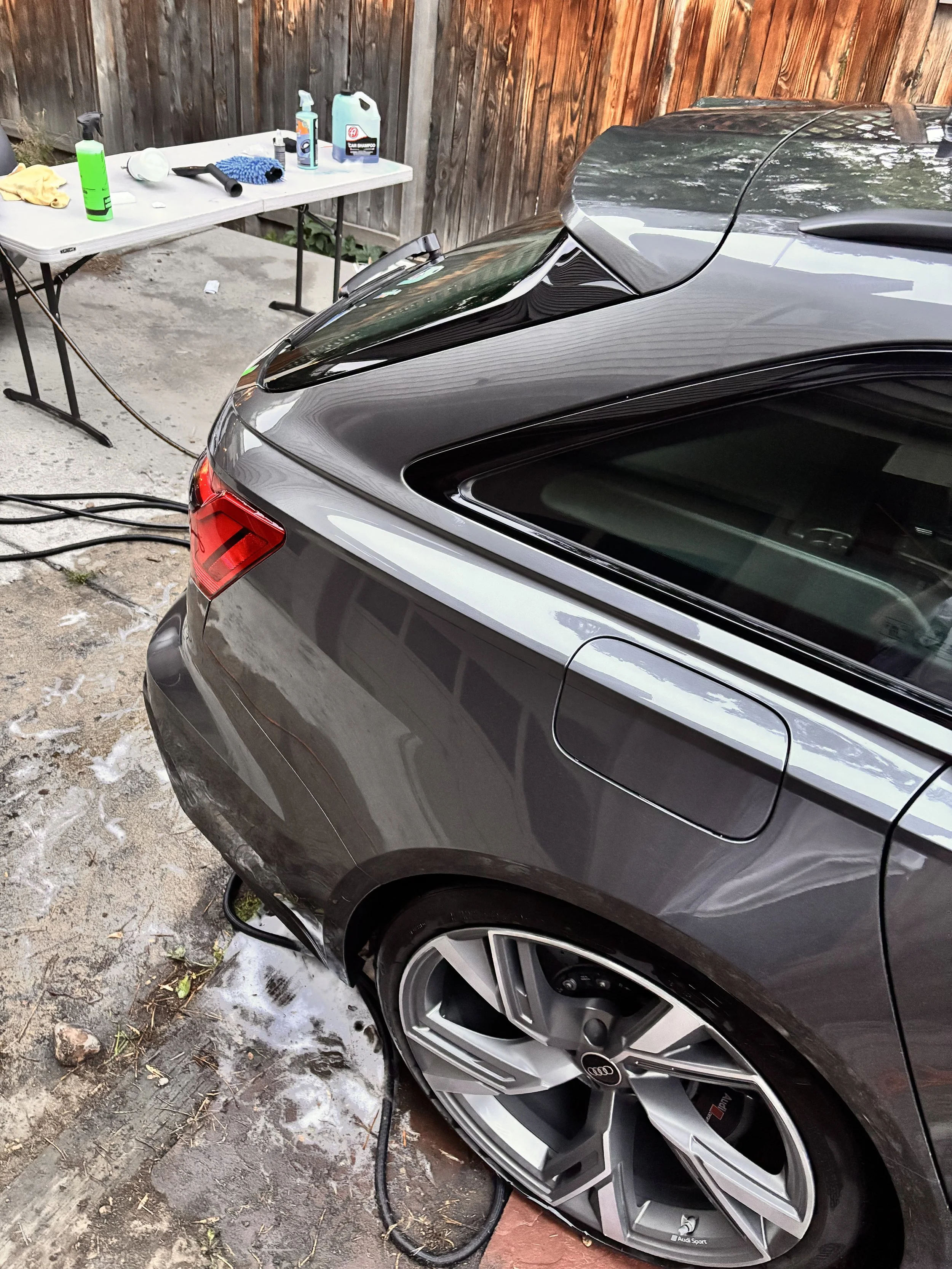 Close-up of a black Audi parked outdoors during a car wash, with washing supplies and a table in the background.